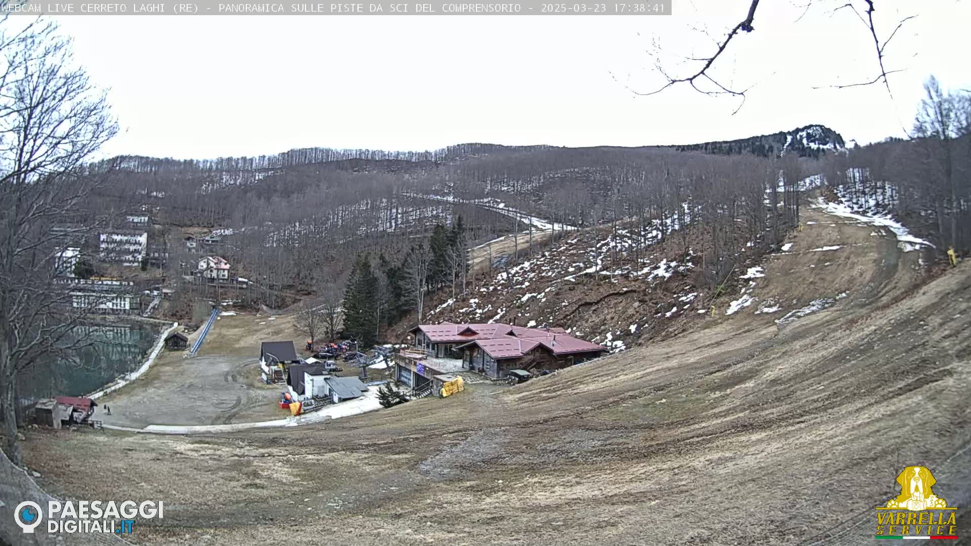A mostly barren ski slope with a building at its base, overlooking a village and lake under a cloudy sky.
