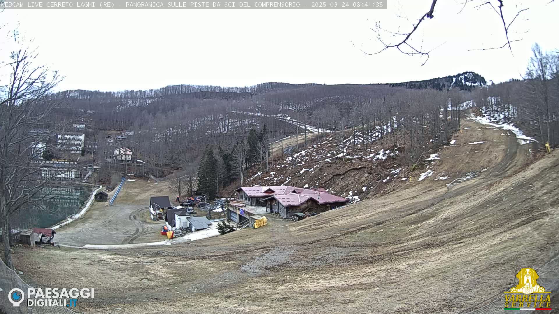 A mostly barren ski slope with a building at its base is situated amongst leafless trees on a cloudy day.