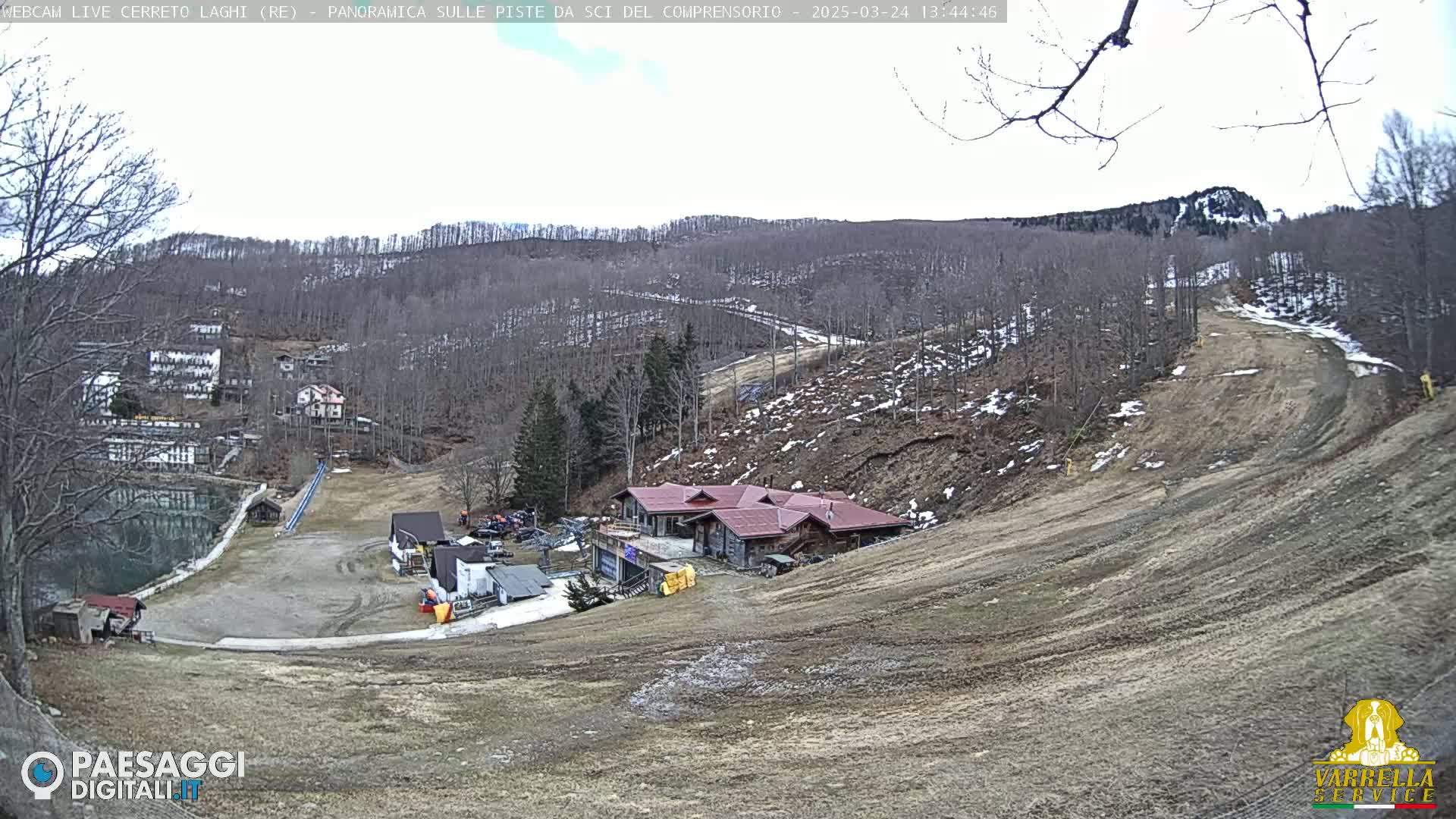 A mostly barren ski slope with a building at its base, surrounded by leafless trees and patches of snow under a mostly cloudy sky.