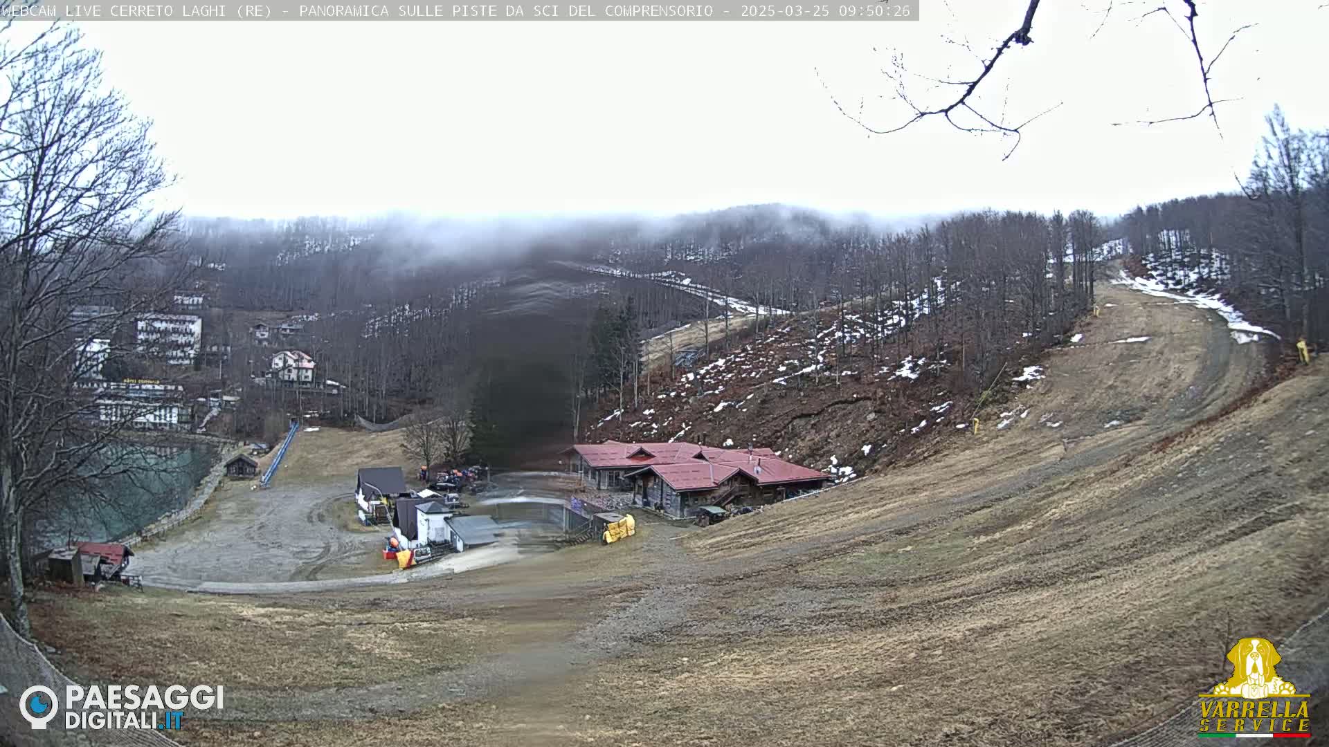 A mostly cloudy, misty day at a ski resort shows a brown, barren ski slope leading down to a cluster of buildings near a body of water and a village nestled into a hillside.