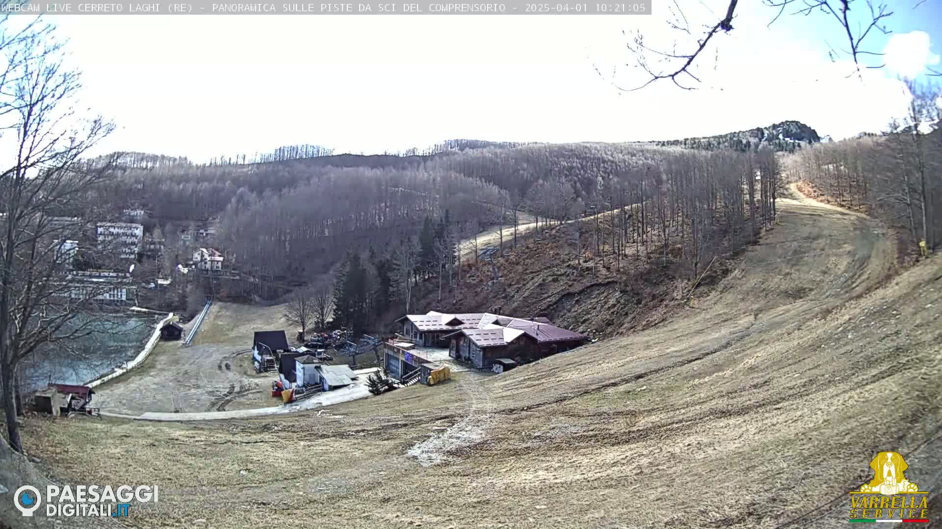 A mostly barren ski slope with a building at the bottom, surrounded by leafless trees under a partly sunny sky.