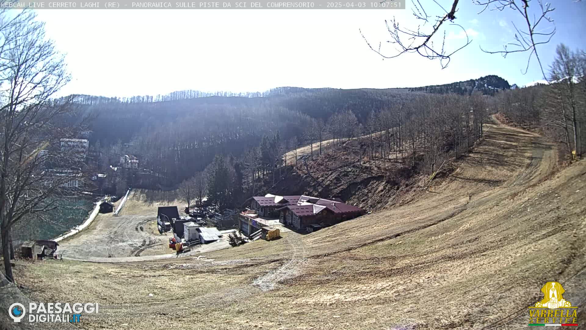 A sunny view of a ski slope with a building at the bottom, barren trees in the background, and a mostly clear sky.