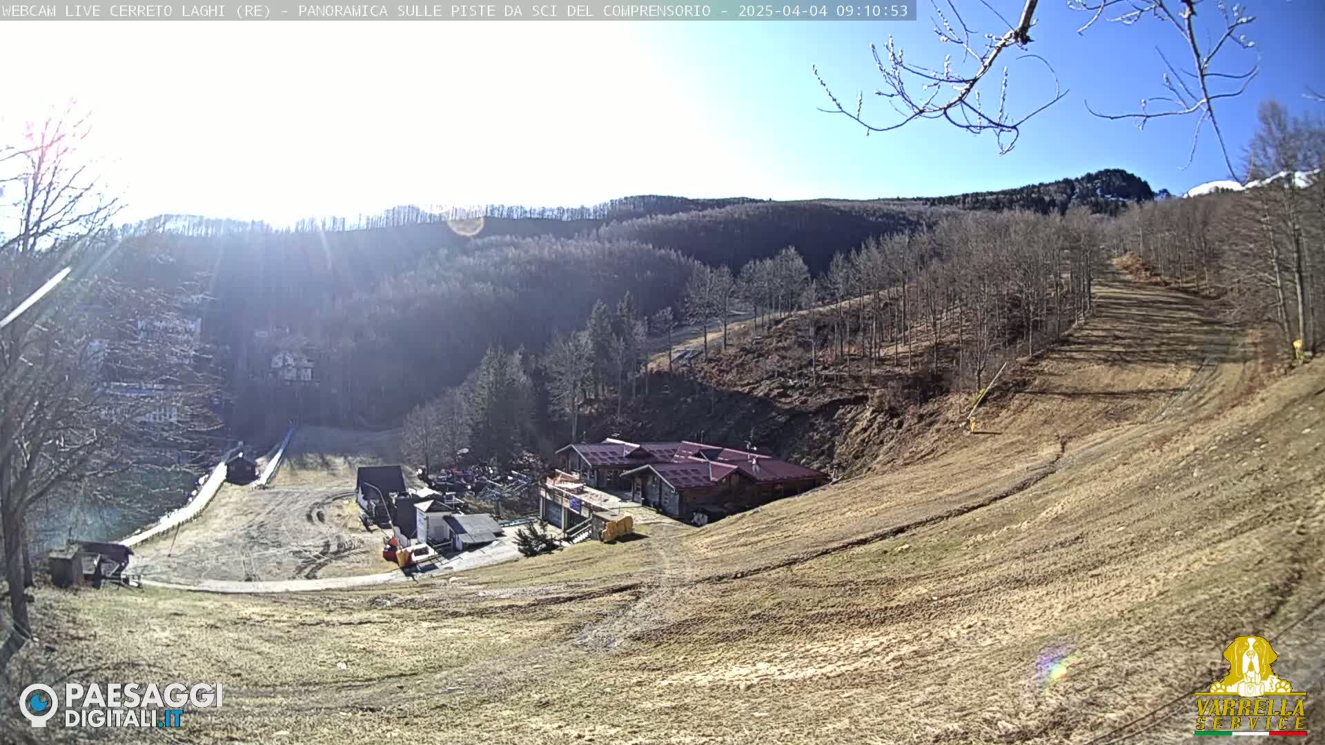 A sunny, high-angle view of a ski slope with a building and surrounding bare trees.