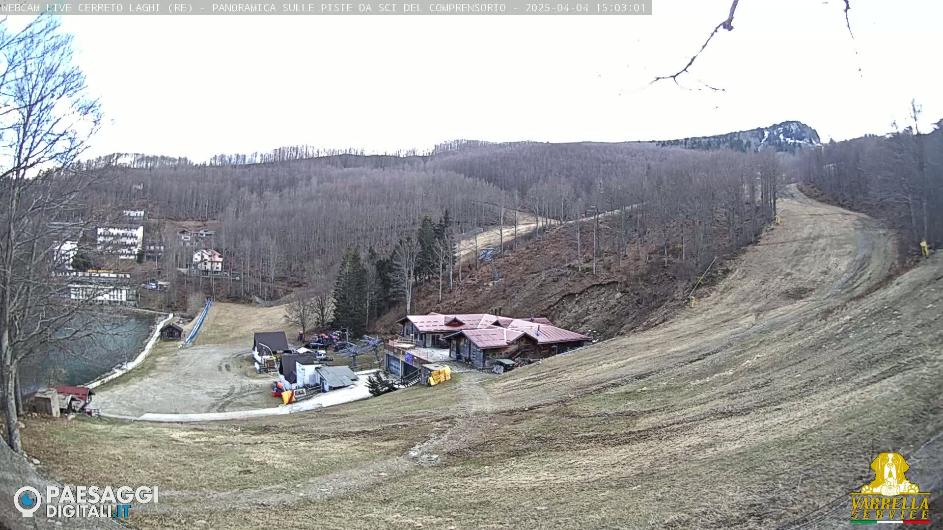A mostly barren ski slope with a building and a small lake at the bottom of a hill on an overcast day.