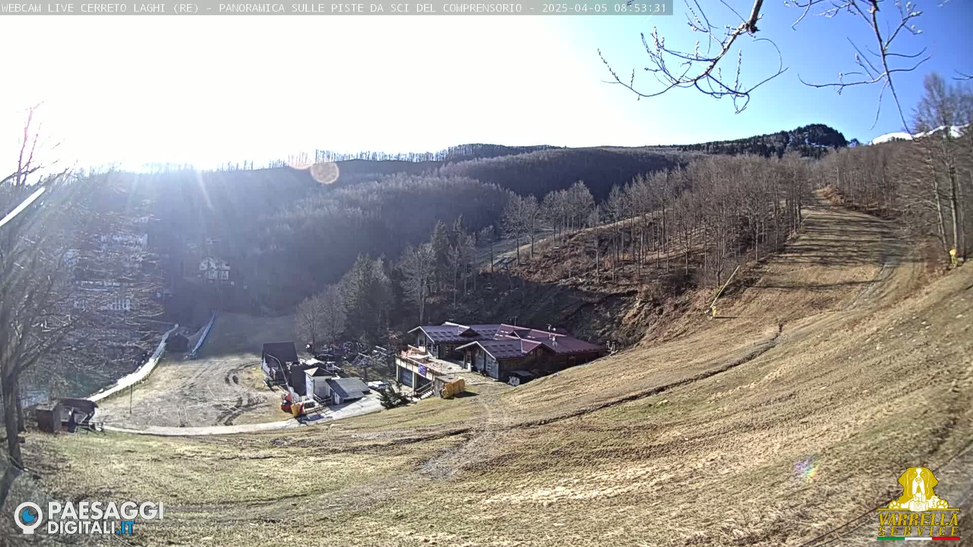 A sunny day overlooking a ski slope with a building at the bottom of a hill and bare trees on the surrounding hills.