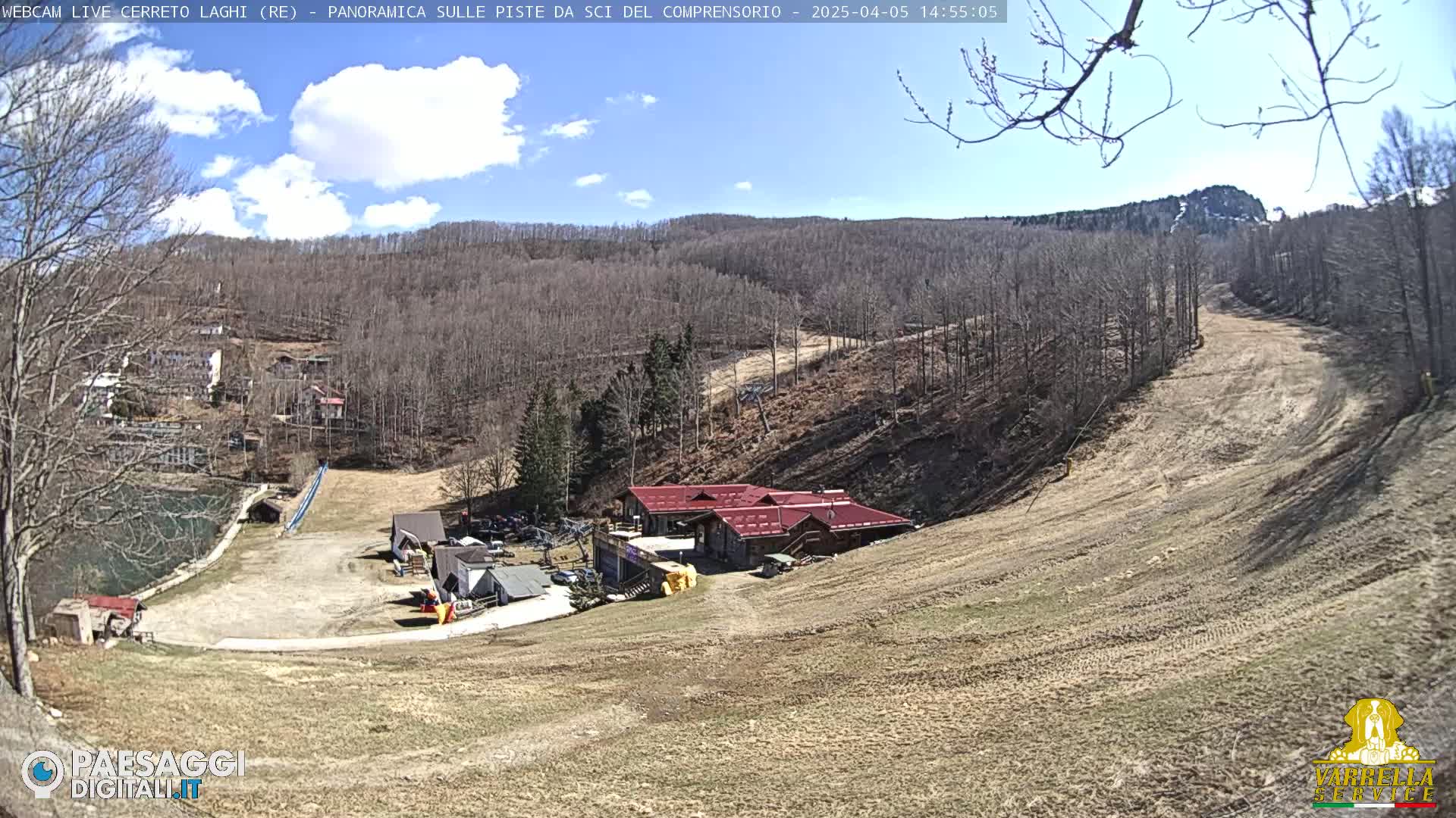 A mostly sunny day reveals a ski slope with a lodge at its base, surrounded by bare trees and brown hills.