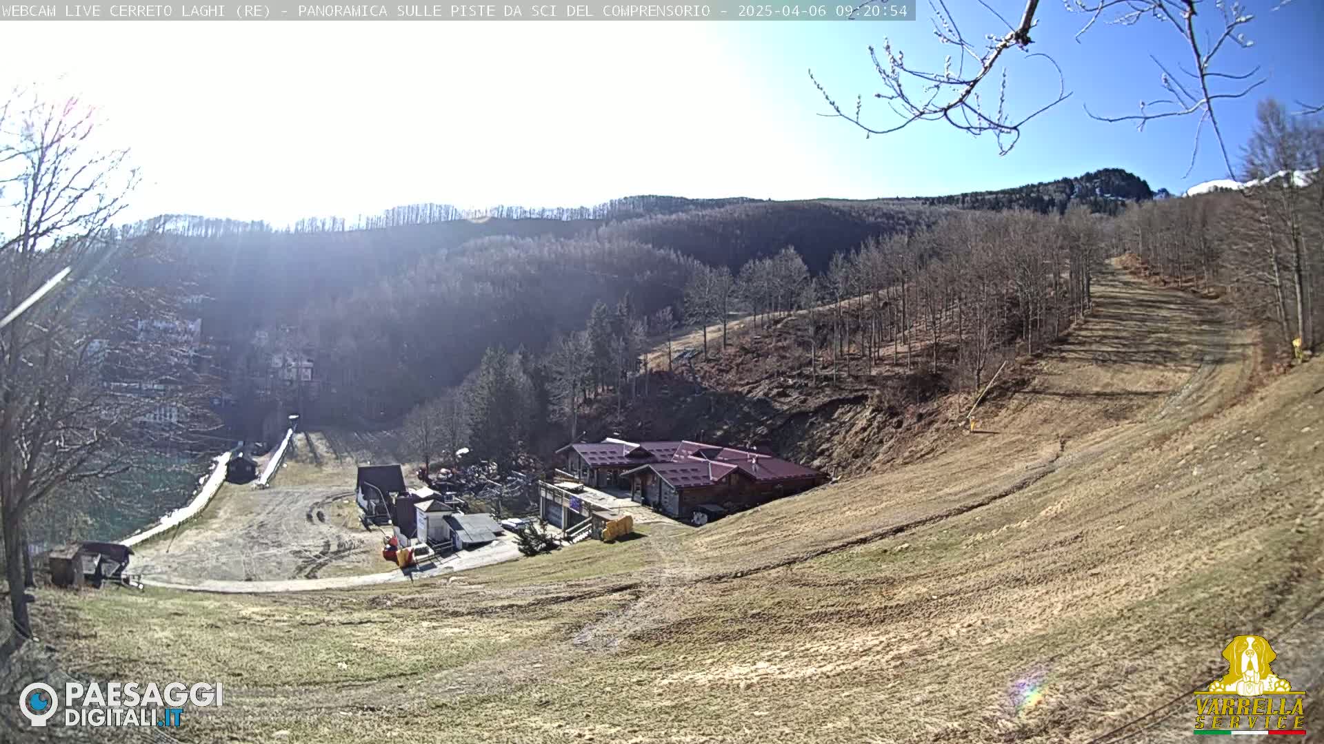 A sunny view of a ski resort with a building at the bottom of a slope, surrounded by leafless trees and brown grass.