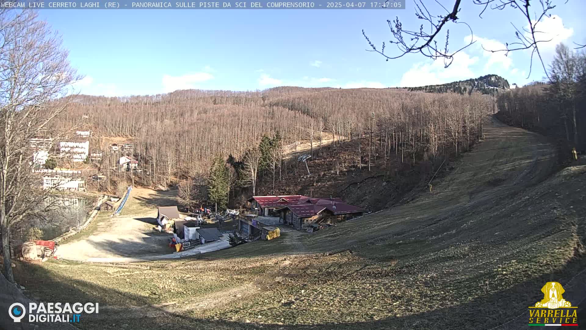 A sunny, mostly clear day reveals a ski slope with a building at its base, surrounded by bare trees on a hillside.
