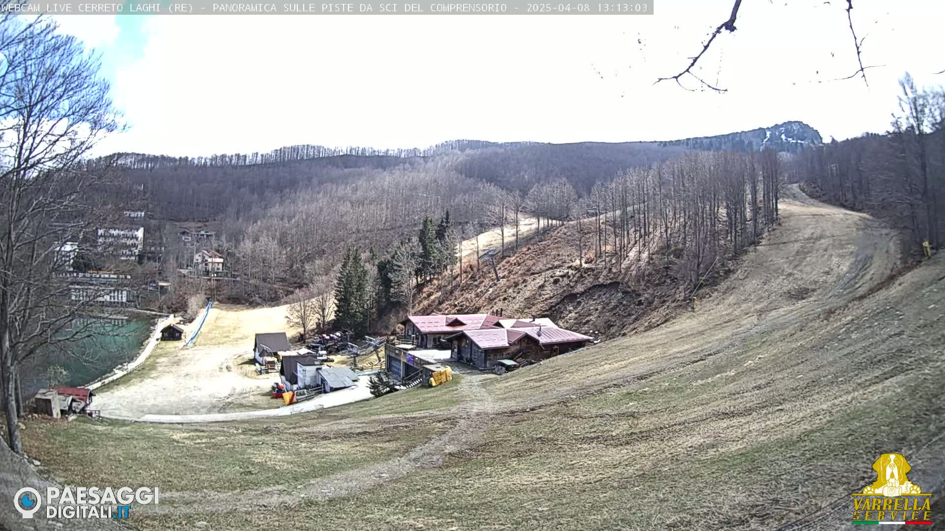 A mostly clear day reveals a ski resort nestled in a brown, leafless forest on a hillside, with a small lake visible in the distance.