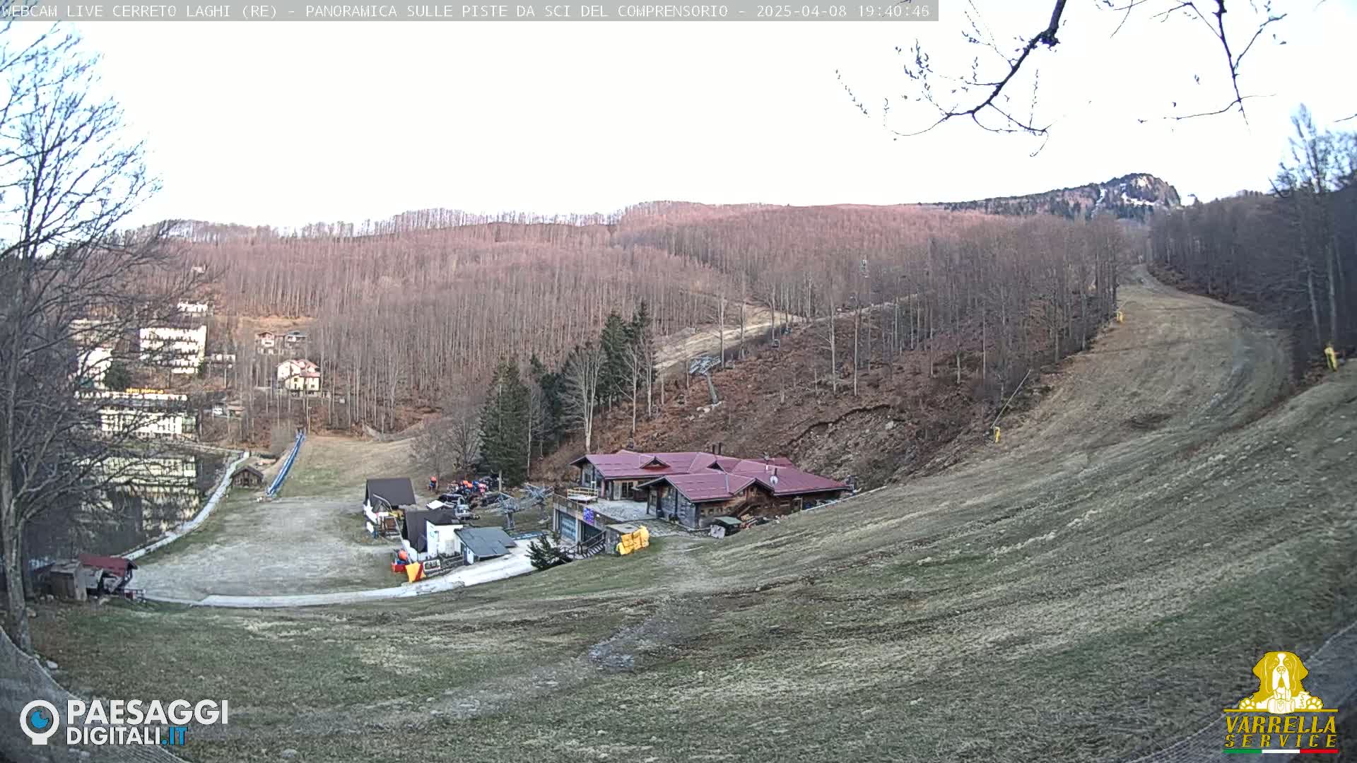 A high-angle, long shot of a ski slope with a building near the bottom, surrounded by bare trees on a clear day.