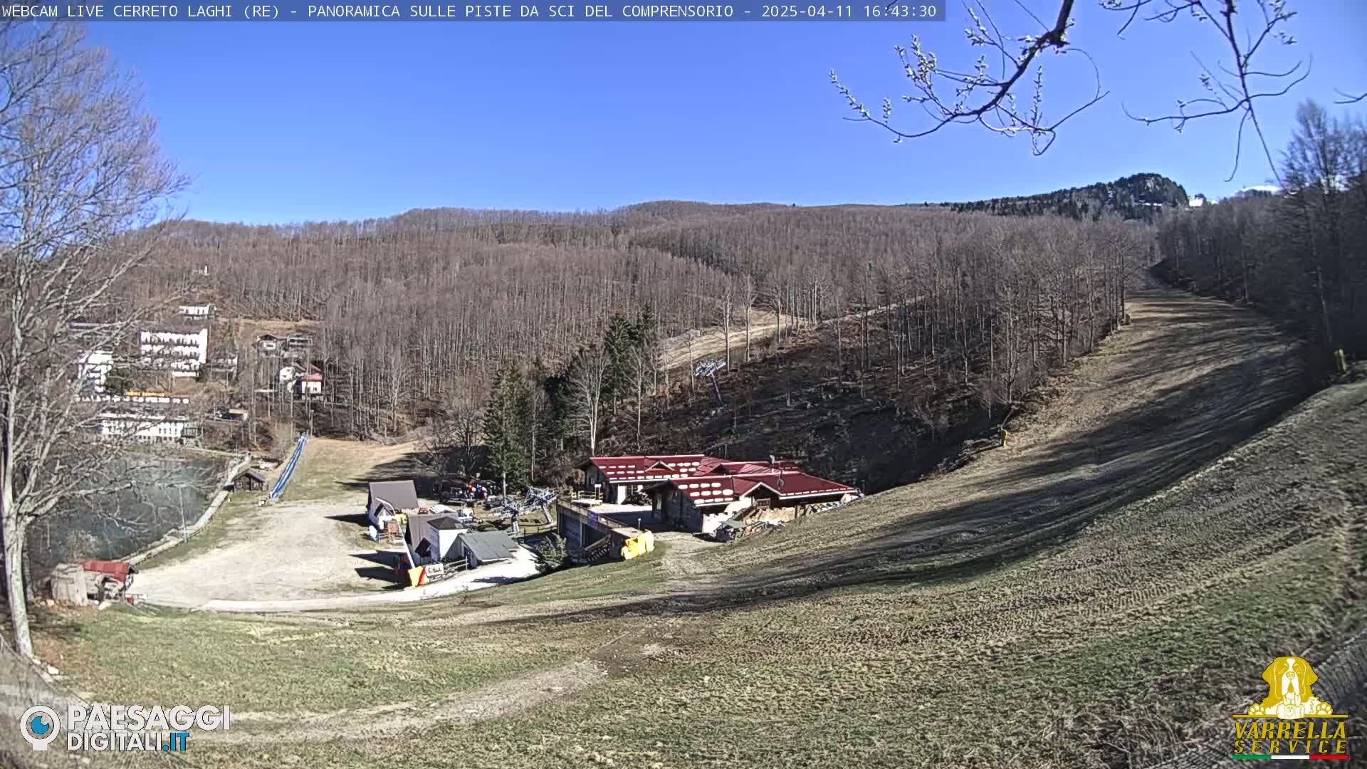 A sunny day overlooking a ski resort with mostly bare trees, a small building, and a long, empty ski slope.