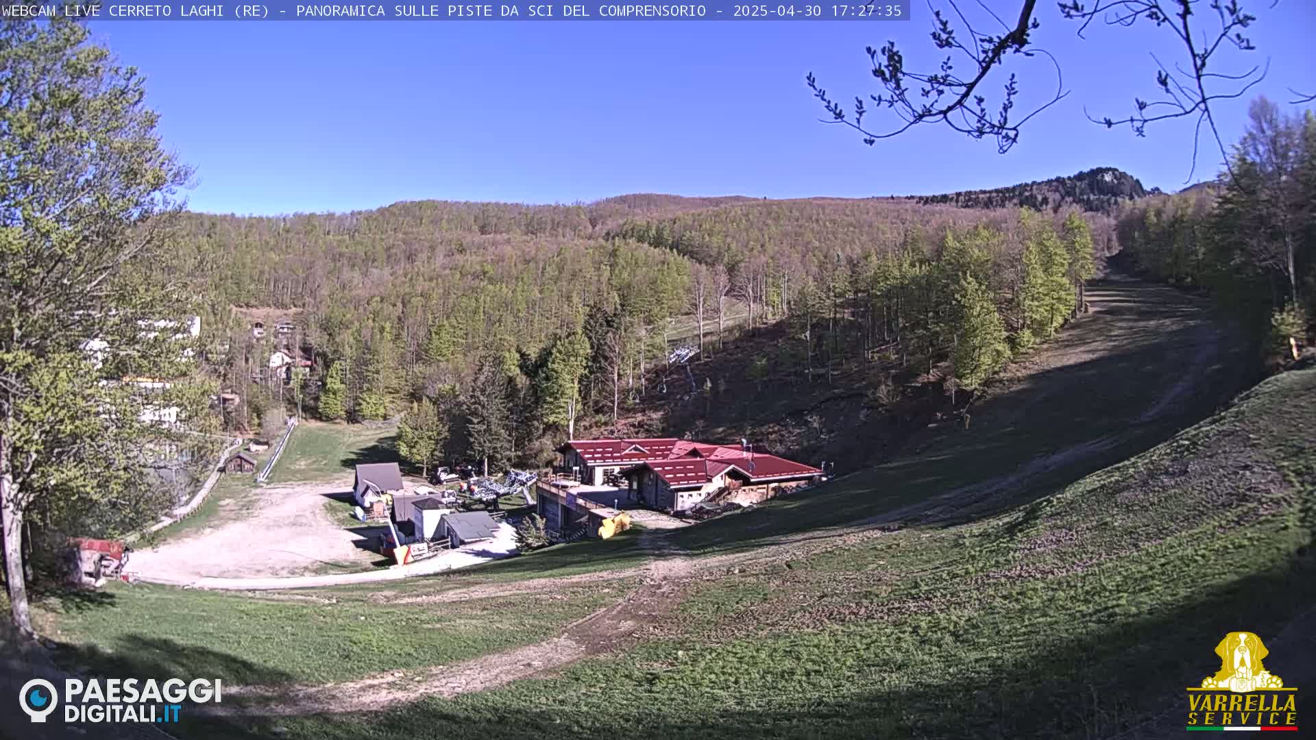 A sunny view of a ski resort in the mountains, showing a small building and grassy ski slopes.