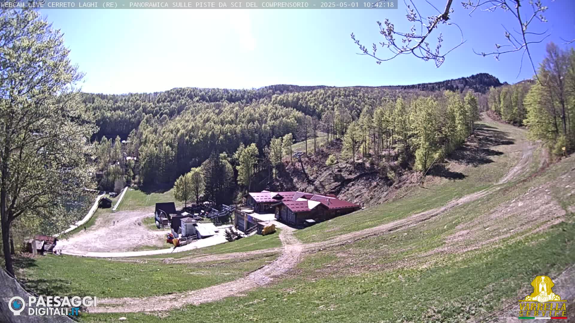 A mountainside ski resort with a red-roofed building is visible under a clear, sunny sky, surrounded by green hills and trees.