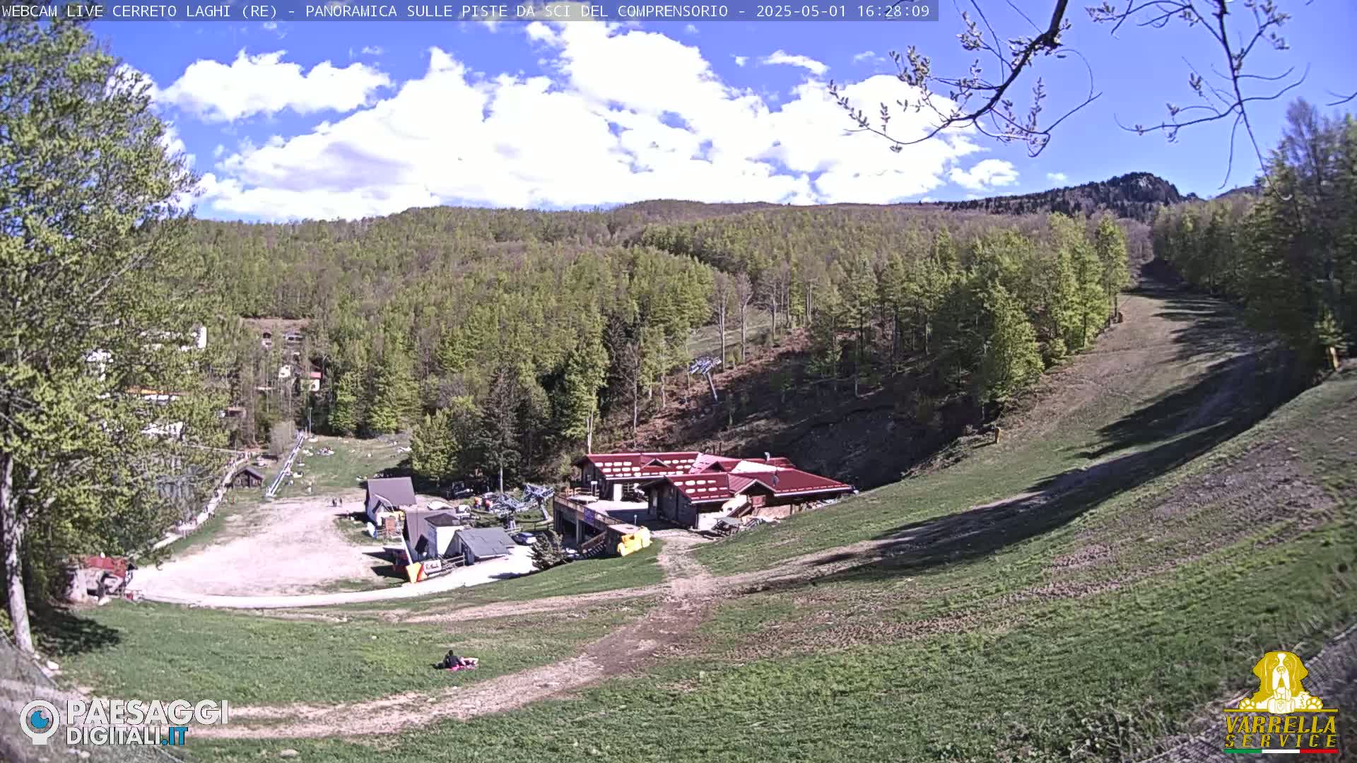 A mostly sunny day reveals a ski resort with a red-roofed building at the base of a hill covered in green trees and grassy slopes.
