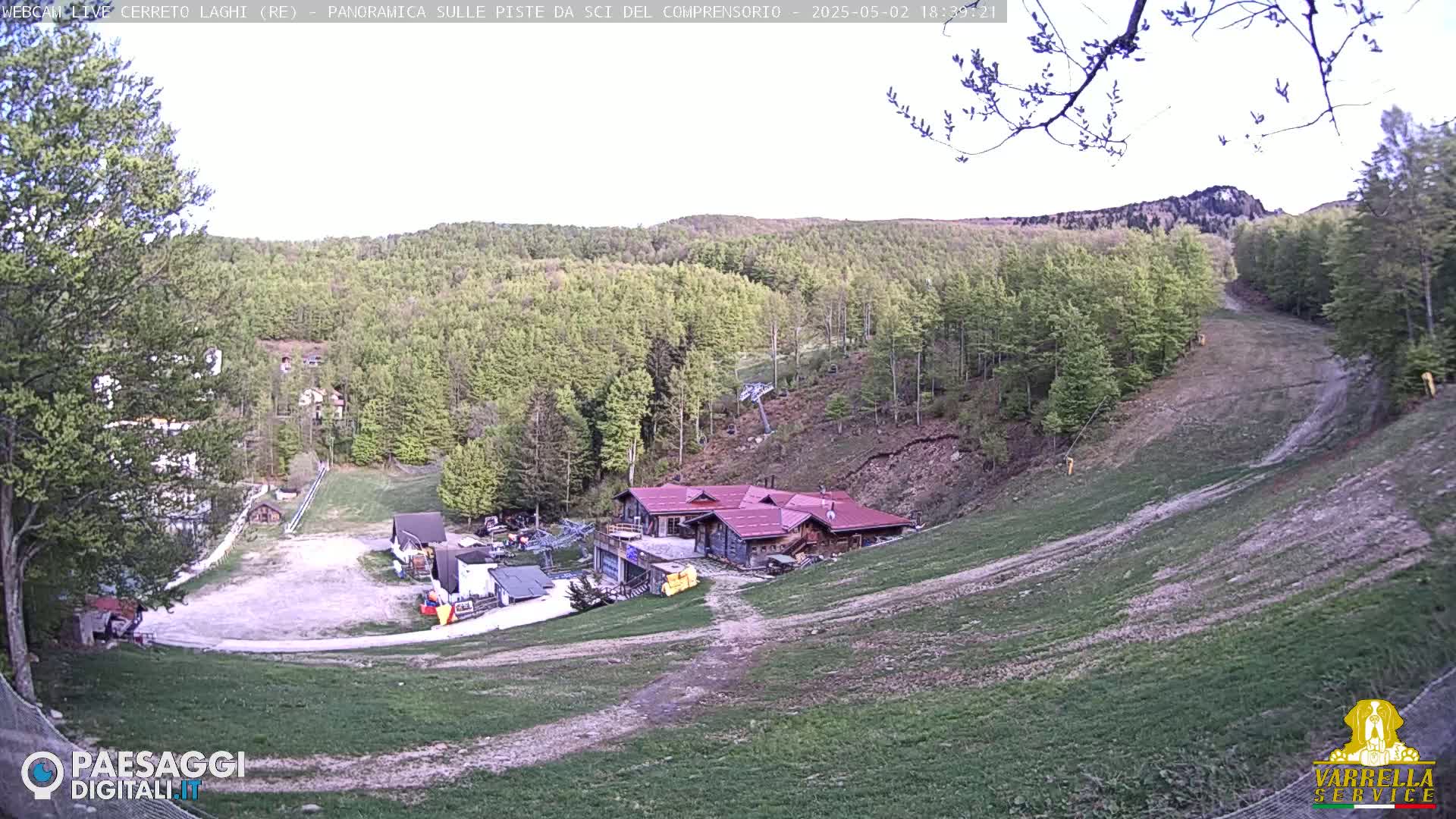 A hillside ski slope with a building at the bottom, surrounded by a forest of light green trees under a mostly clear sky.