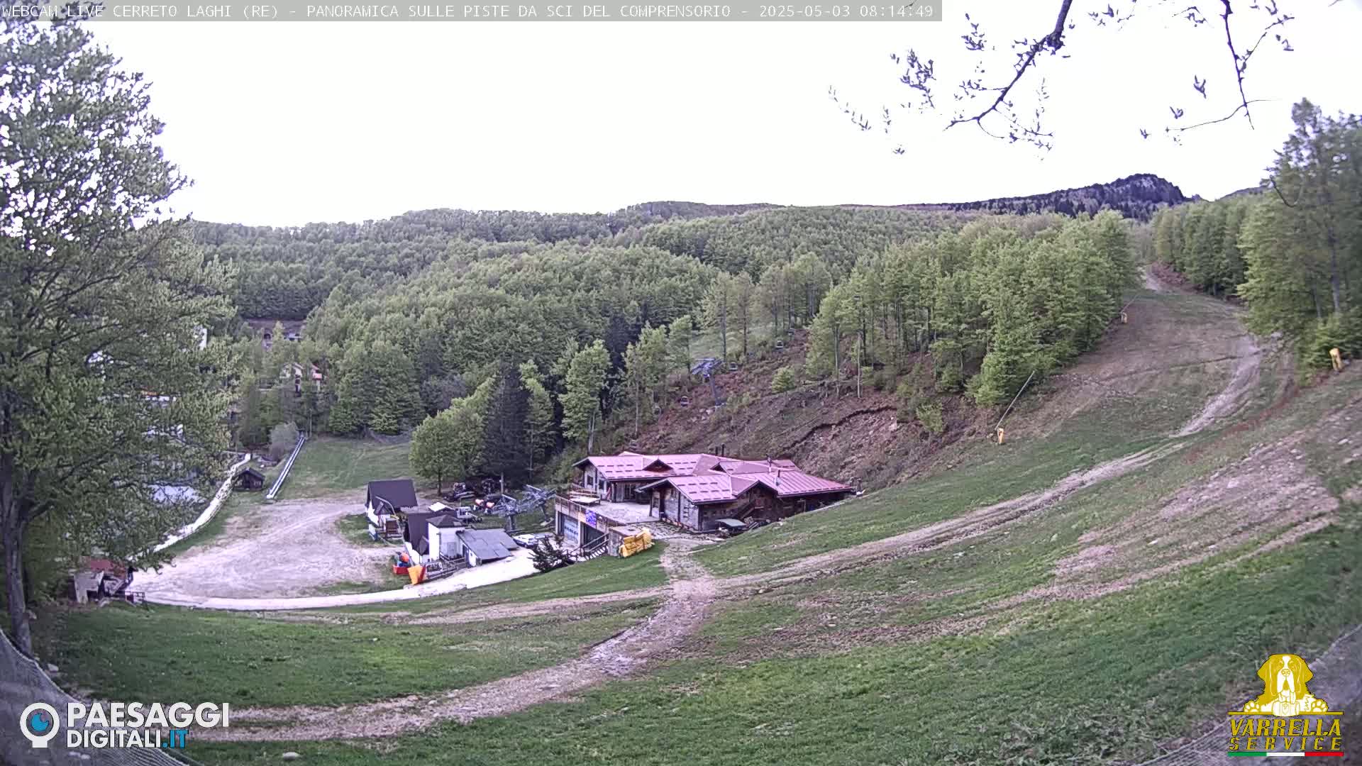 A mostly clear day reveals a ski resort in the hills, with a building near the bottom of a grassy slope, surrounded by lush green trees.