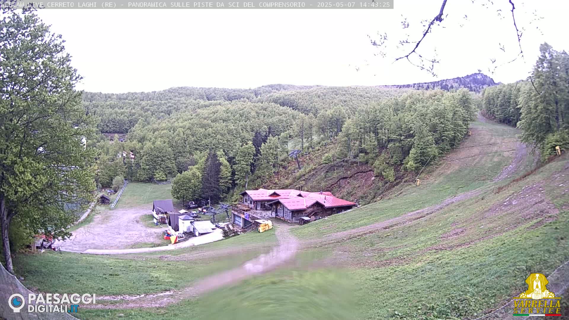 A mostly clear day reveals a ski resort with a red-roofed building at the base of a grassy hill lined with trees, and a ski lift extending upwards.