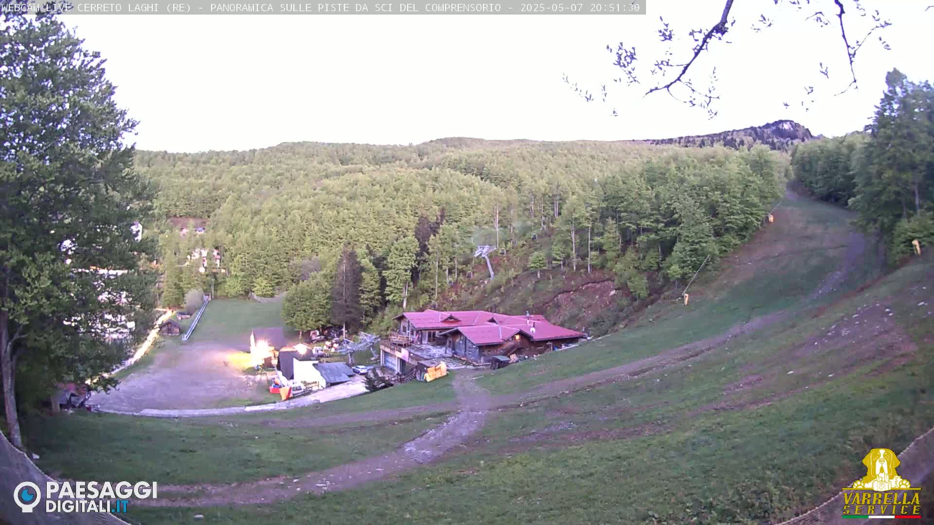A hilltop ski resort area at dusk, showing a building, ski lift, and grassy slopes with a few trees.