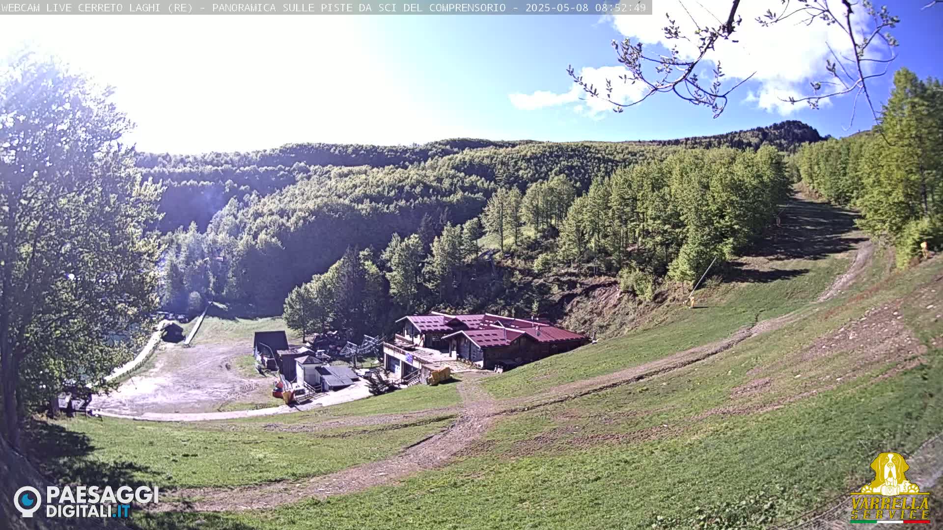 A mostly sunny day reveals a mountainside ski resort with a building complex at the base of a grassy slope, surrounded by green trees under a partly cloudy sky.