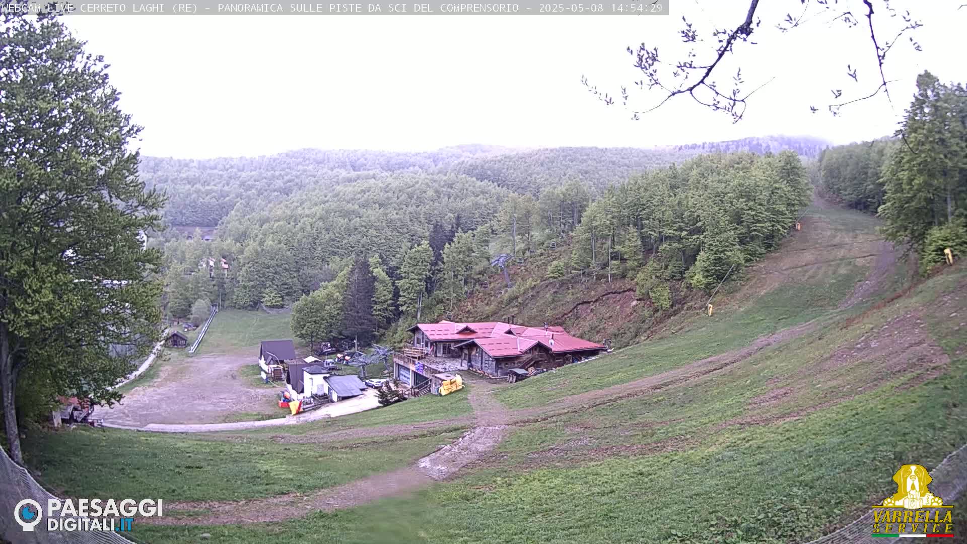 A mostly overcast view of a ski resort in the springtime, showing a building at the base of a grassy hill surrounded by lush green deciduous forests.