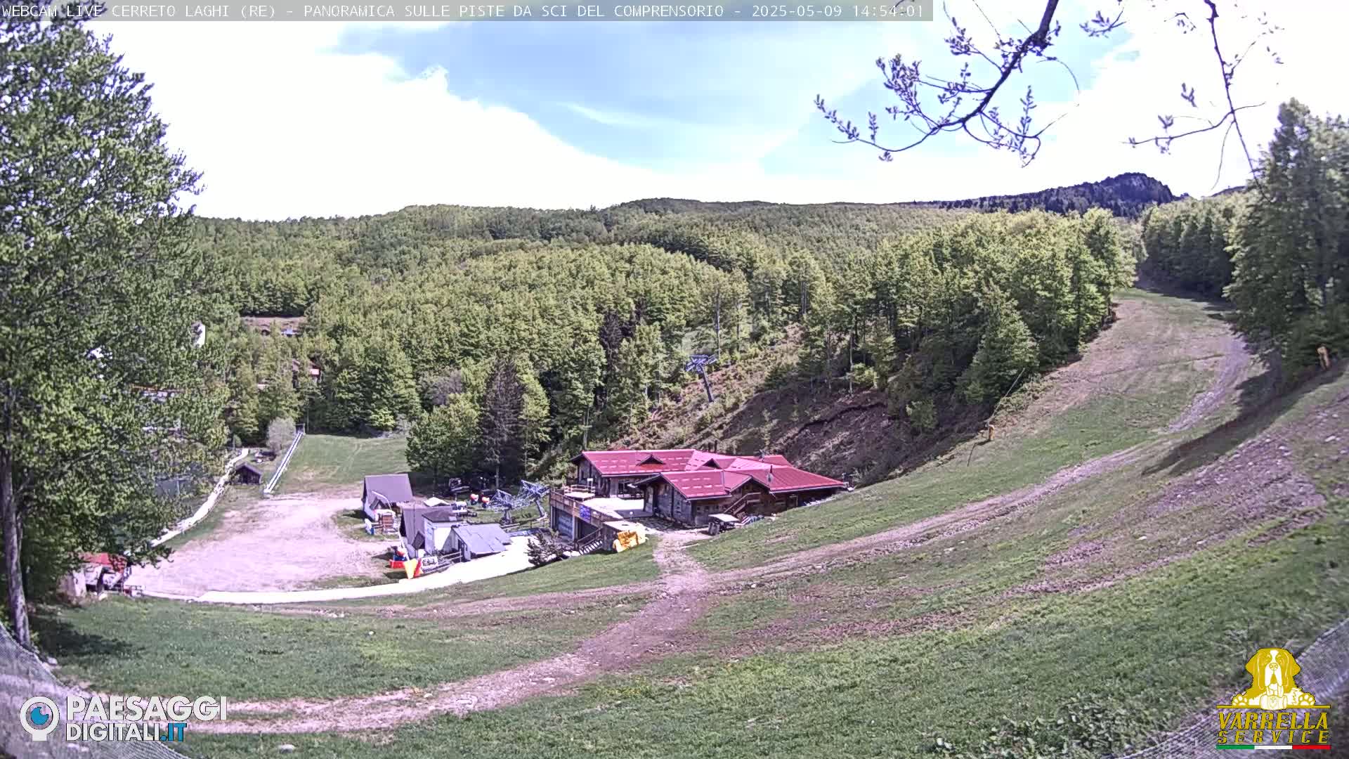 A mostly sunny day reveals a ski resort in a green, hilly landscape with a red-roofed building near the bottom of a grassy slope.