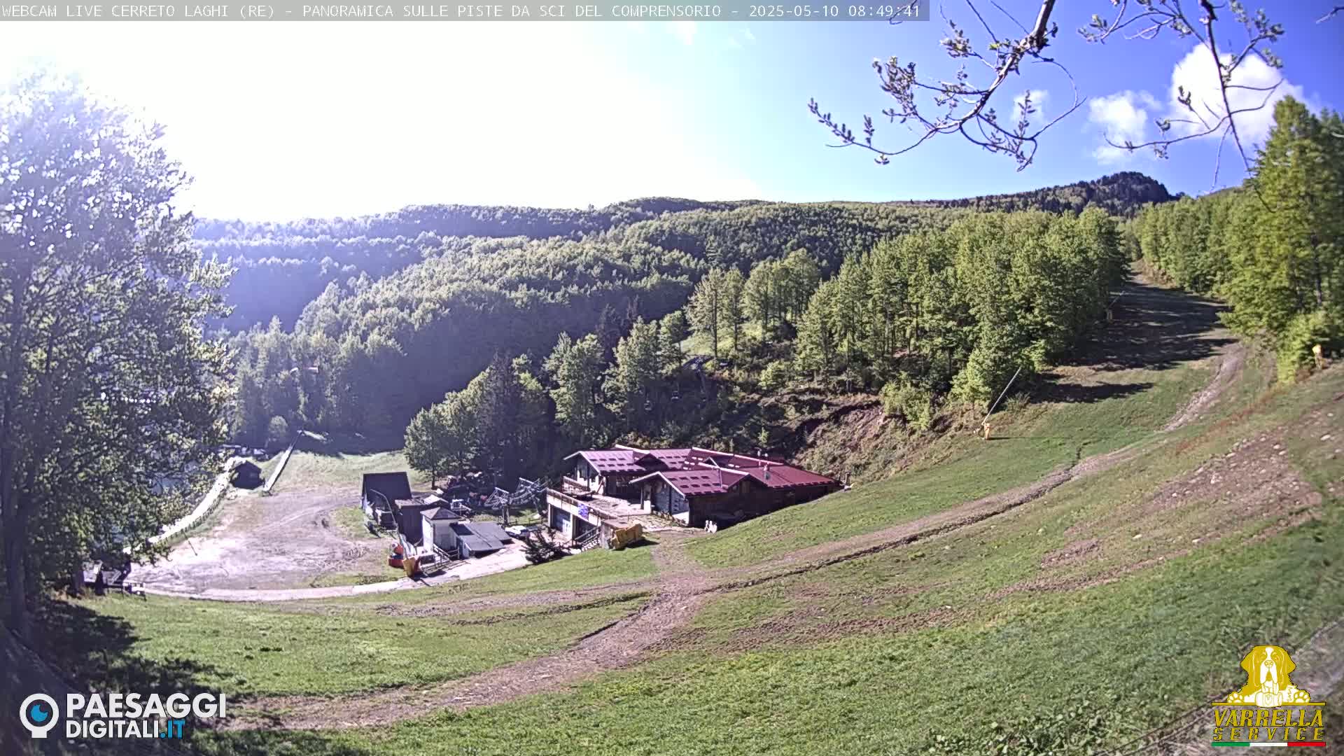 A mountainside ski resort with a building and grassy slopes is shown under a mostly sunny sky.