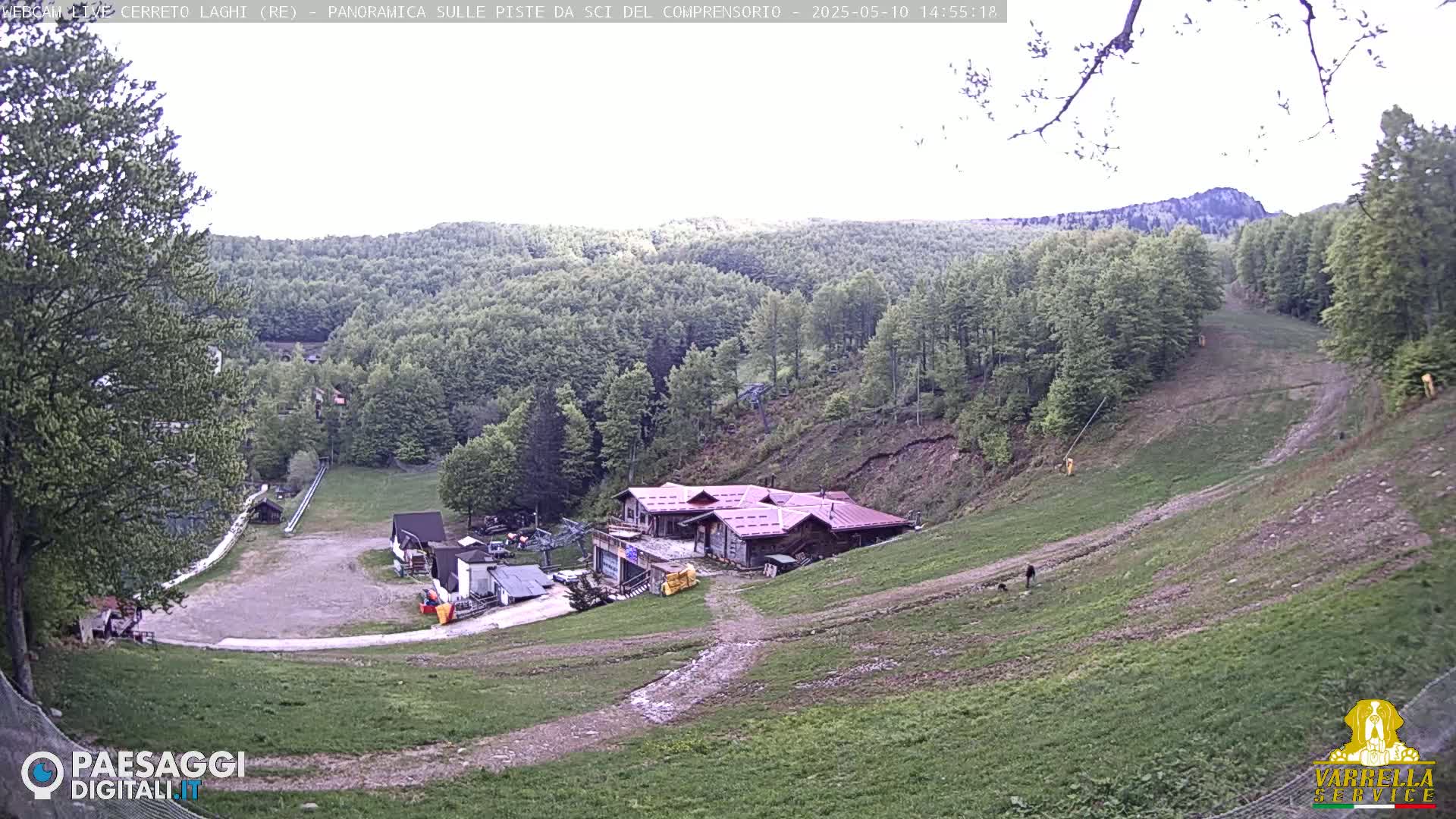 A mostly clear day reveals a ski slope with a building at the bottom, surrounded by green hills and trees.