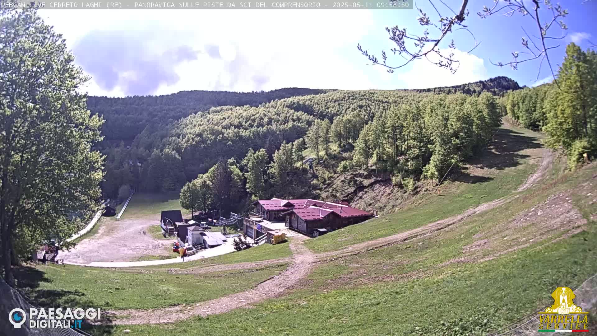 A mostly sunny view of a ski resort in the mountains, with grassy slopes and a building at the base.