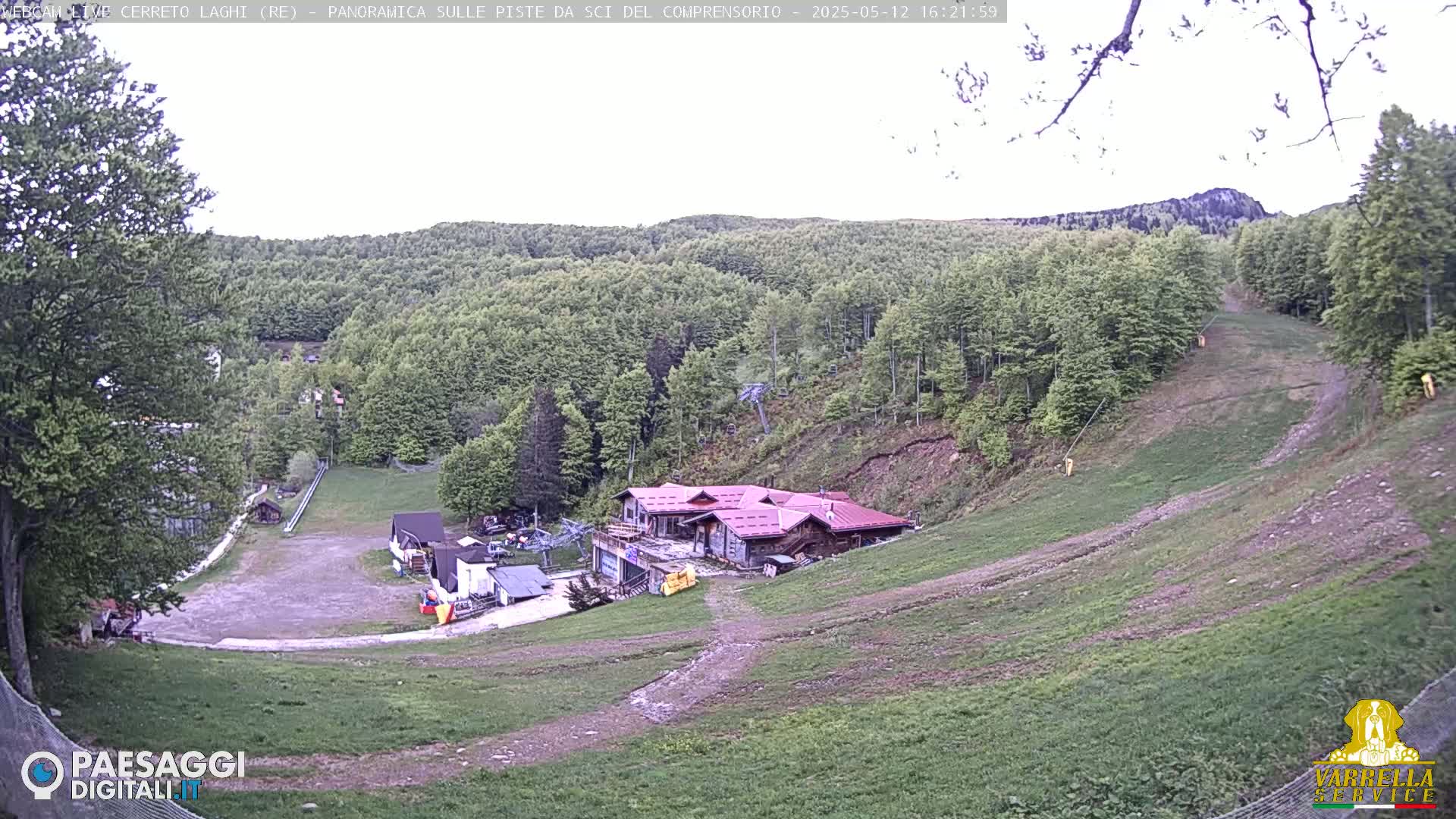 A mostly clear, daytime view of a ski resort in a wooded area, showing a building near the bottom of a grassy ski slope that leads up a hillside.
