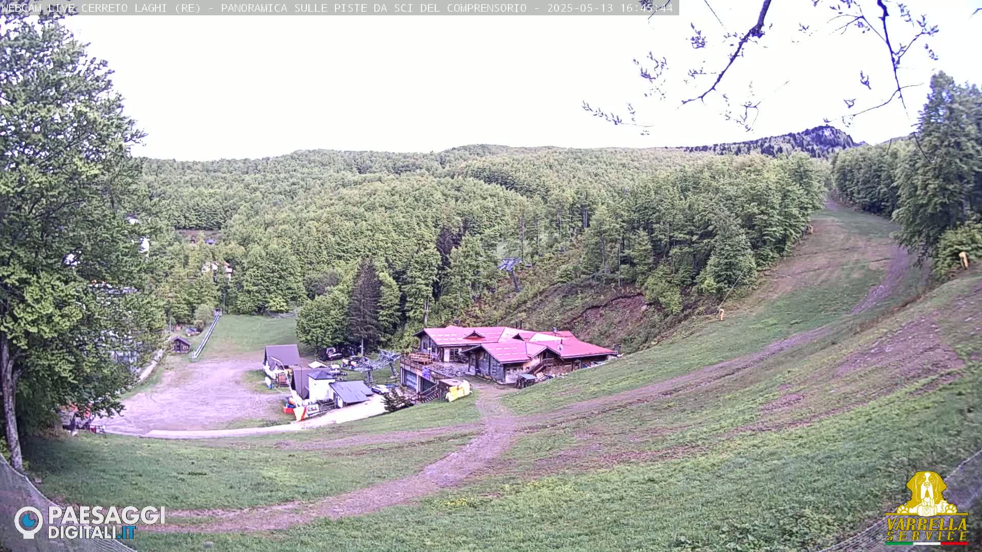 A mostly clear day reveals a grassy hillside with a red-roofed building and several smaller structures near the bottom, all surrounded by lush green trees.