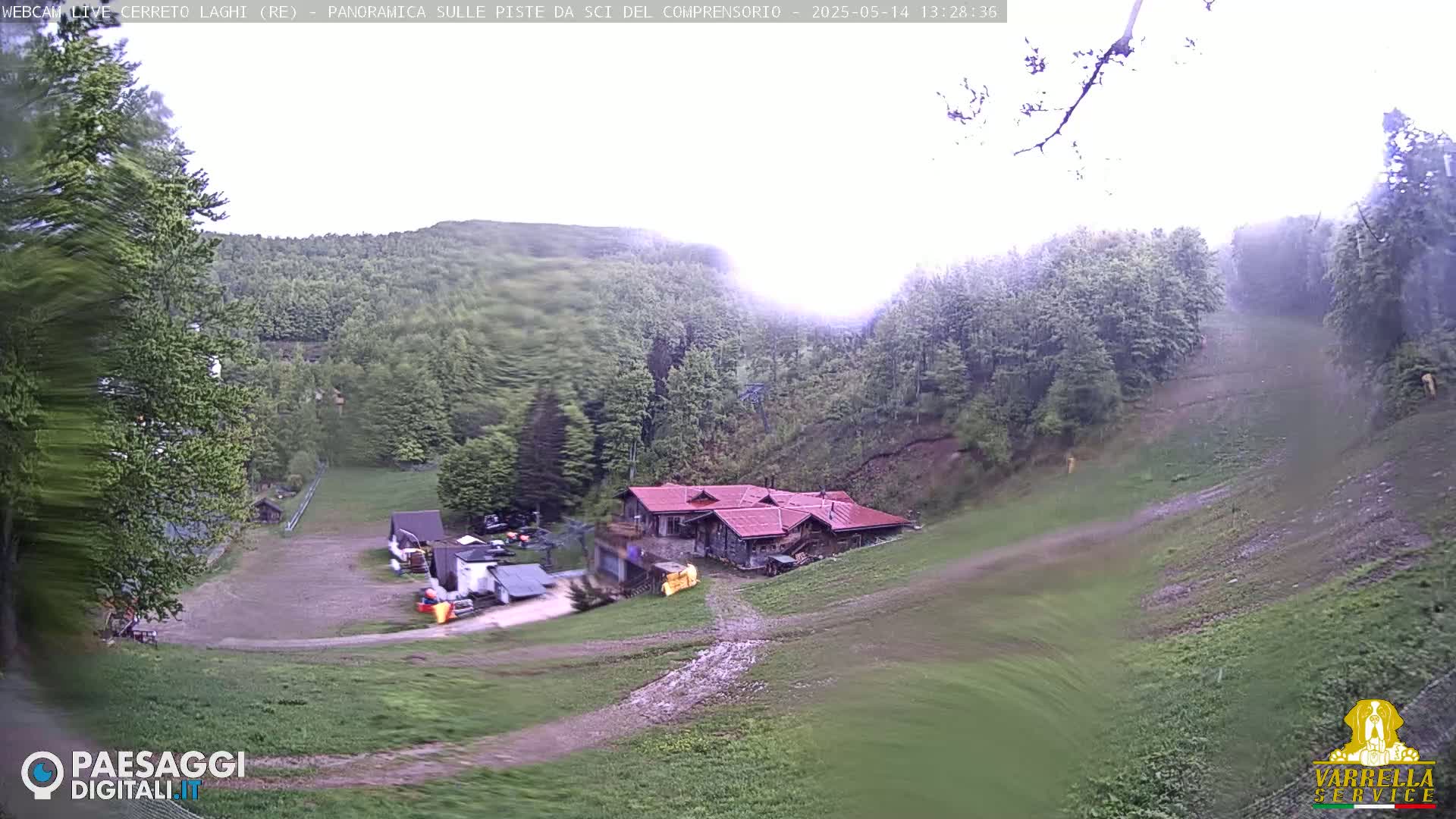 A mountainside view shows a building with a red roof nestled amongst green trees and grassy slopes under an overcast sky.
