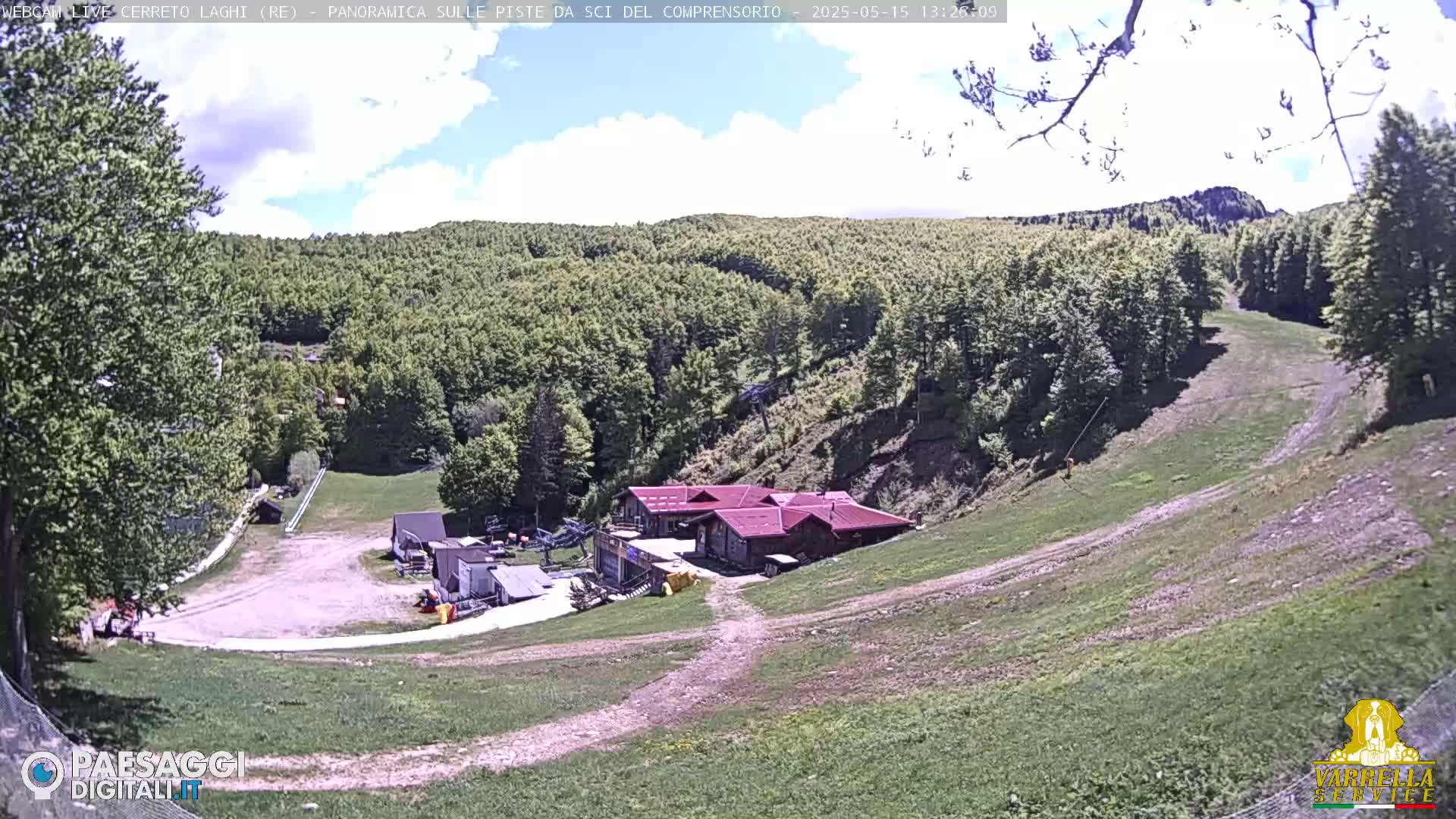 A mostly sunny view of a ski resort in a hilly, wooded area, showing a small cluster of buildings at the base of a grassy ski slope.