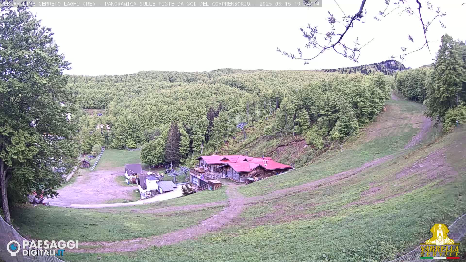 A mostly sunny day reveals a ski resort nestled in a verdant hillside, featuring a large building with a reddish-brown roof and a grassy slope leading down to it.
