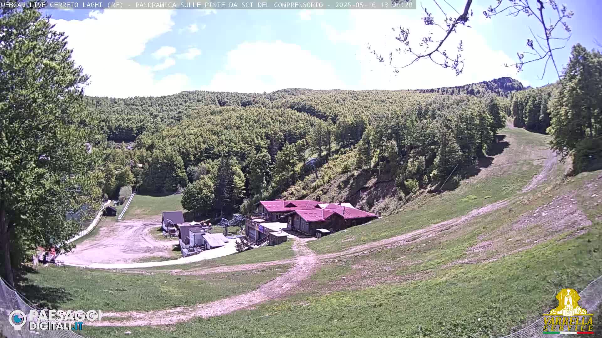 A mostly sunny day reveals a ski slope with a few buildings at its base, surrounded by a lush green forest on a hillside.