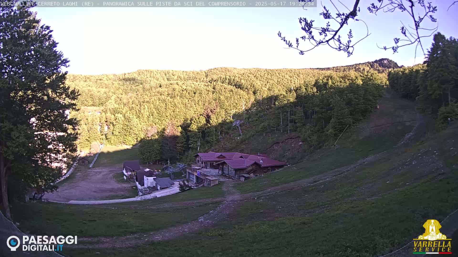 A mountainside ski resort, mostly brown and green, is seen under a clear, sunny sky; a building is centrally located in the foreground.