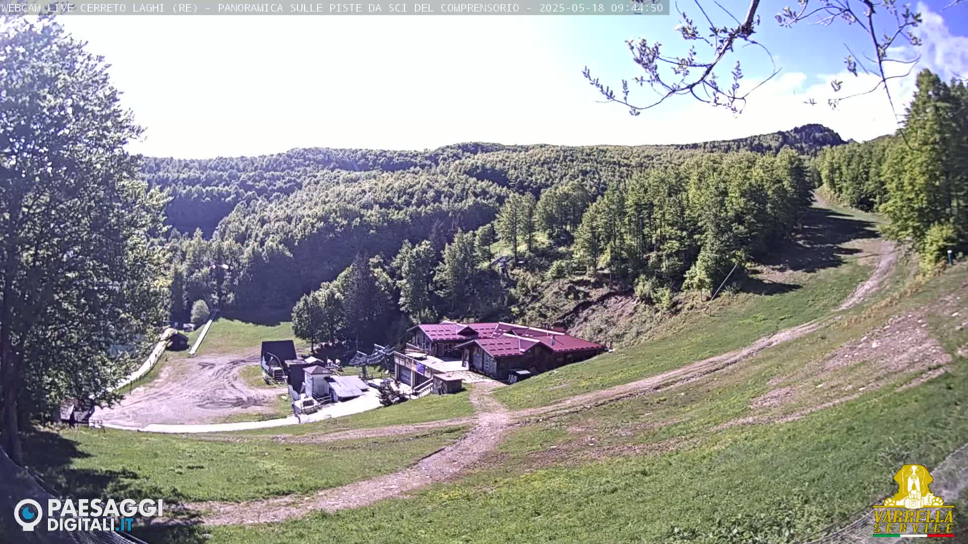 A grassy ski slope with a building at its base, surrounded by green hills and trees under a mostly clear sky.
