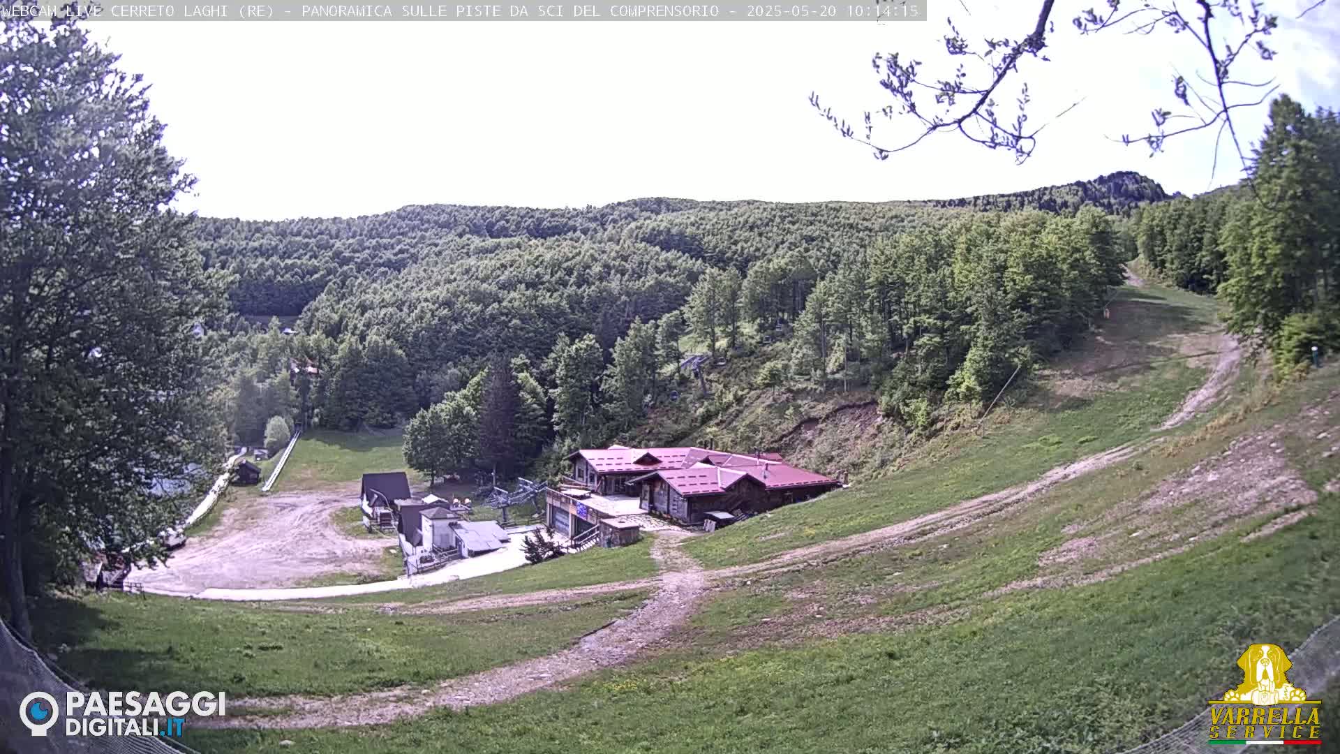 A ski resort in a lush green mountain setting on a partly sunny day shows several buildings at the base of a grassy hill and a mostly tree-covered mountain in the background.