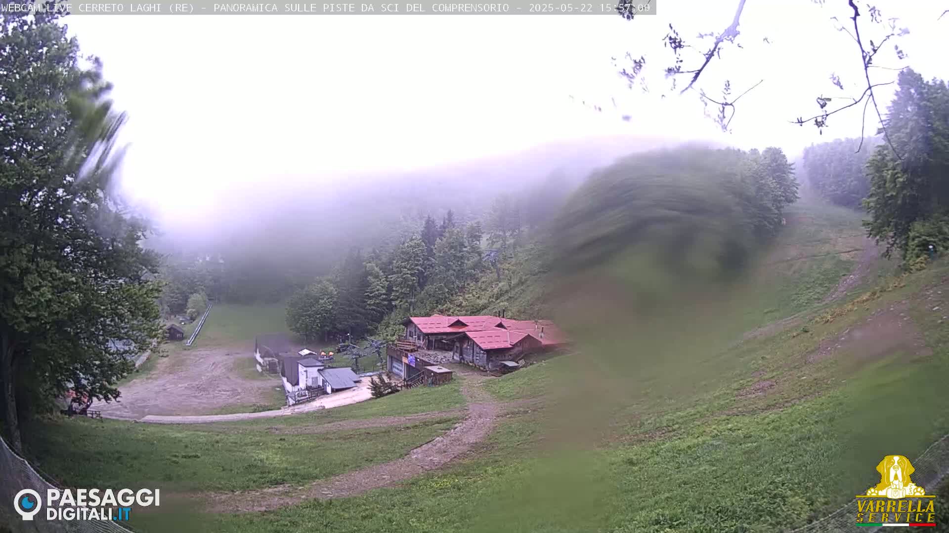 A foggy, windy day overlooks a grassy hillside with several buildings, including one with a red roof, and a ski slope.