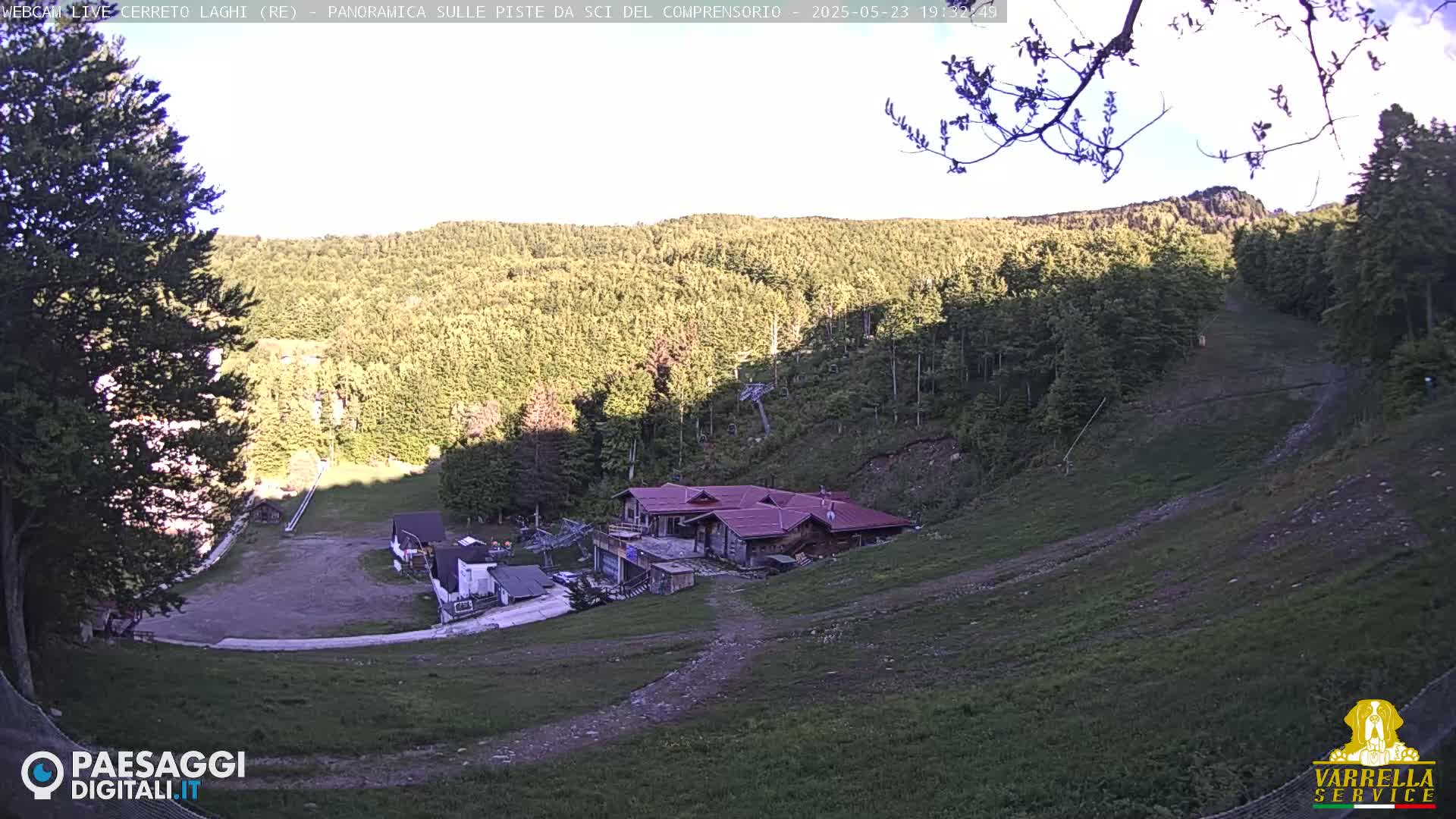 A high-angle view shows a ski resort in the mountains on a partly sunny day, with a building near the bottom of a grassy ski slope surrounded by trees.