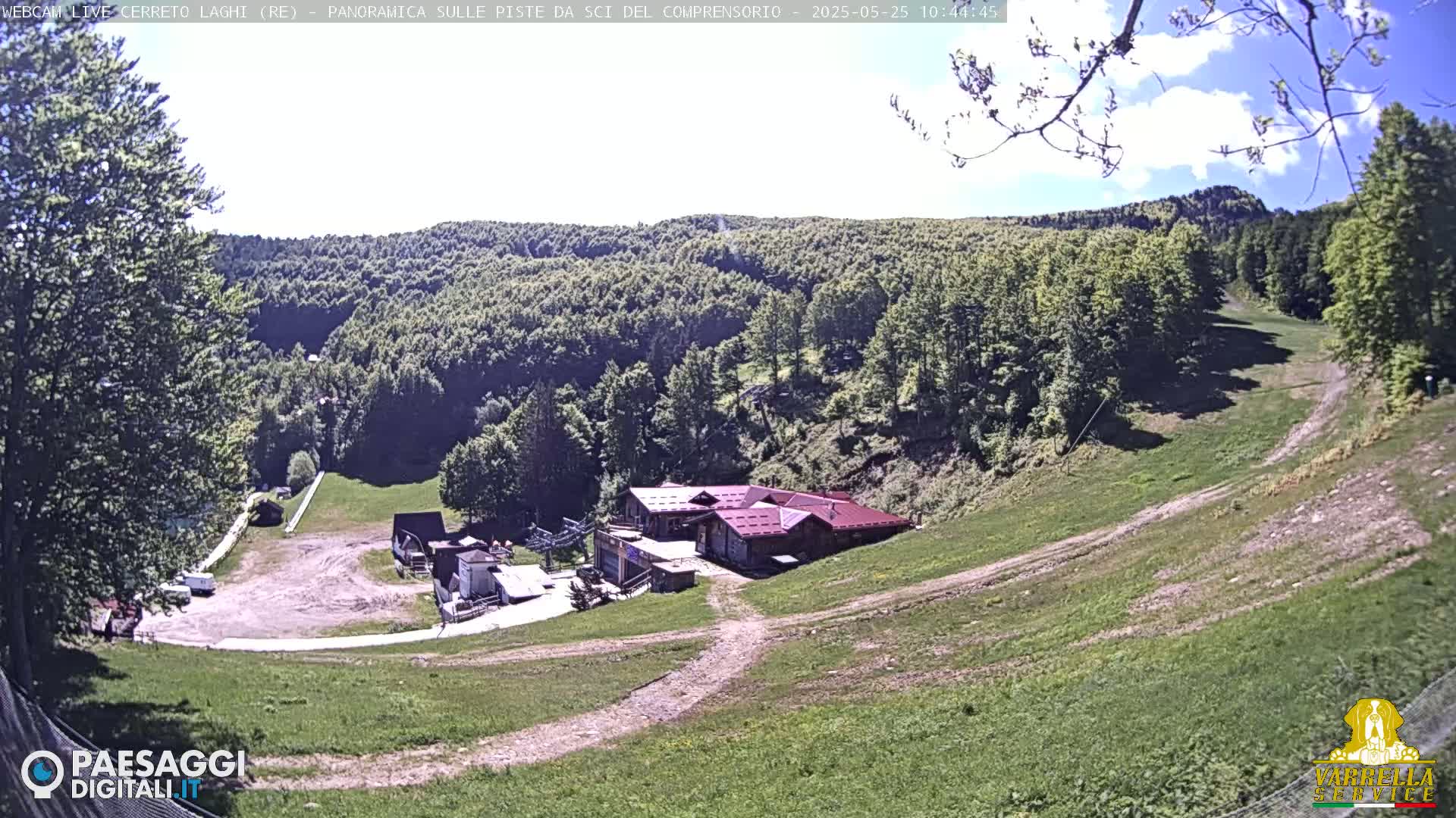 A mostly sunny day overlooking a grassy ski slope with a building and surrounding forest.