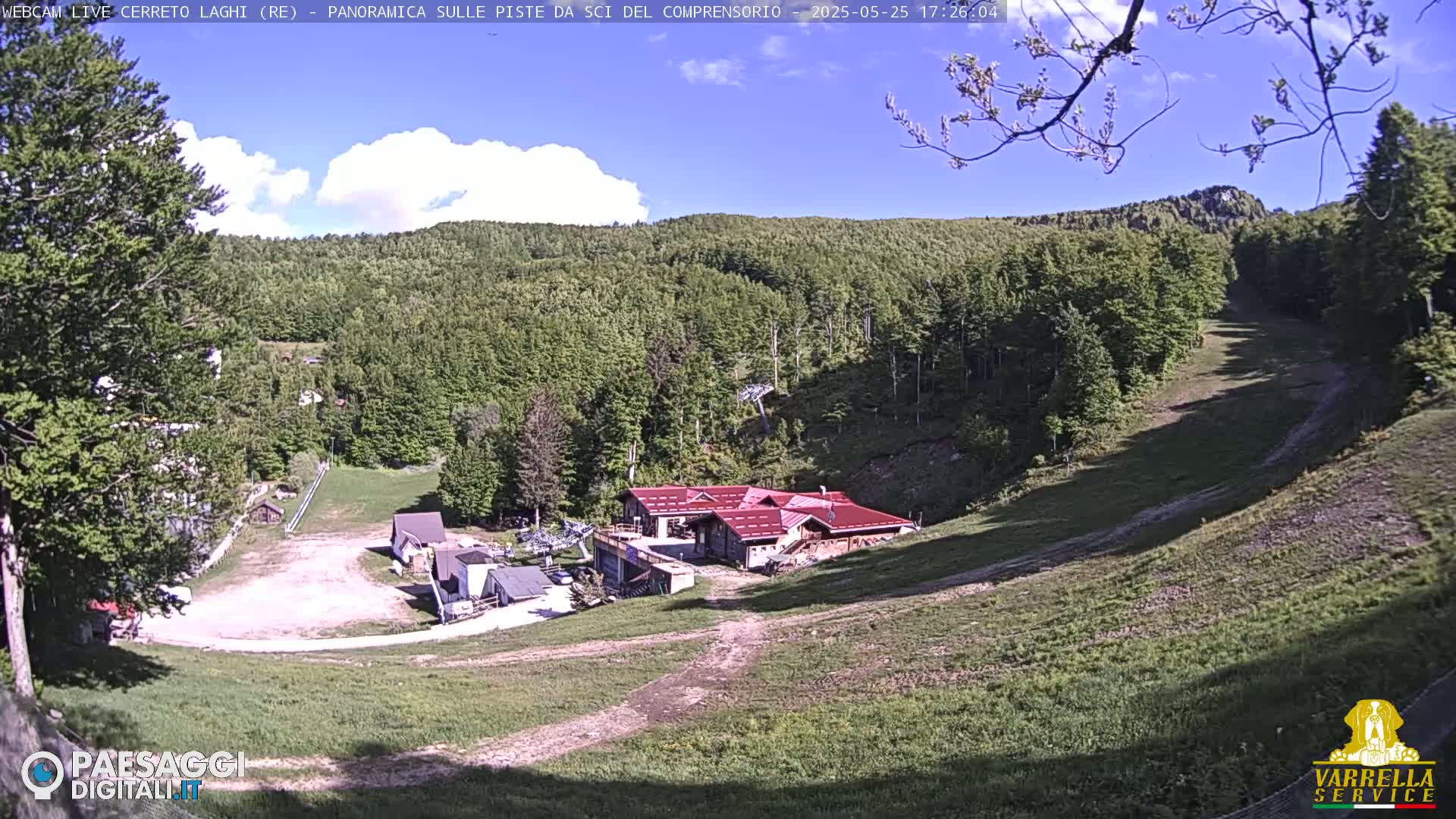 A partly sunny day reveals a ski resort nestled in a verdant hillside, featuring a red-roofed building at the base of a grassy ski slope.