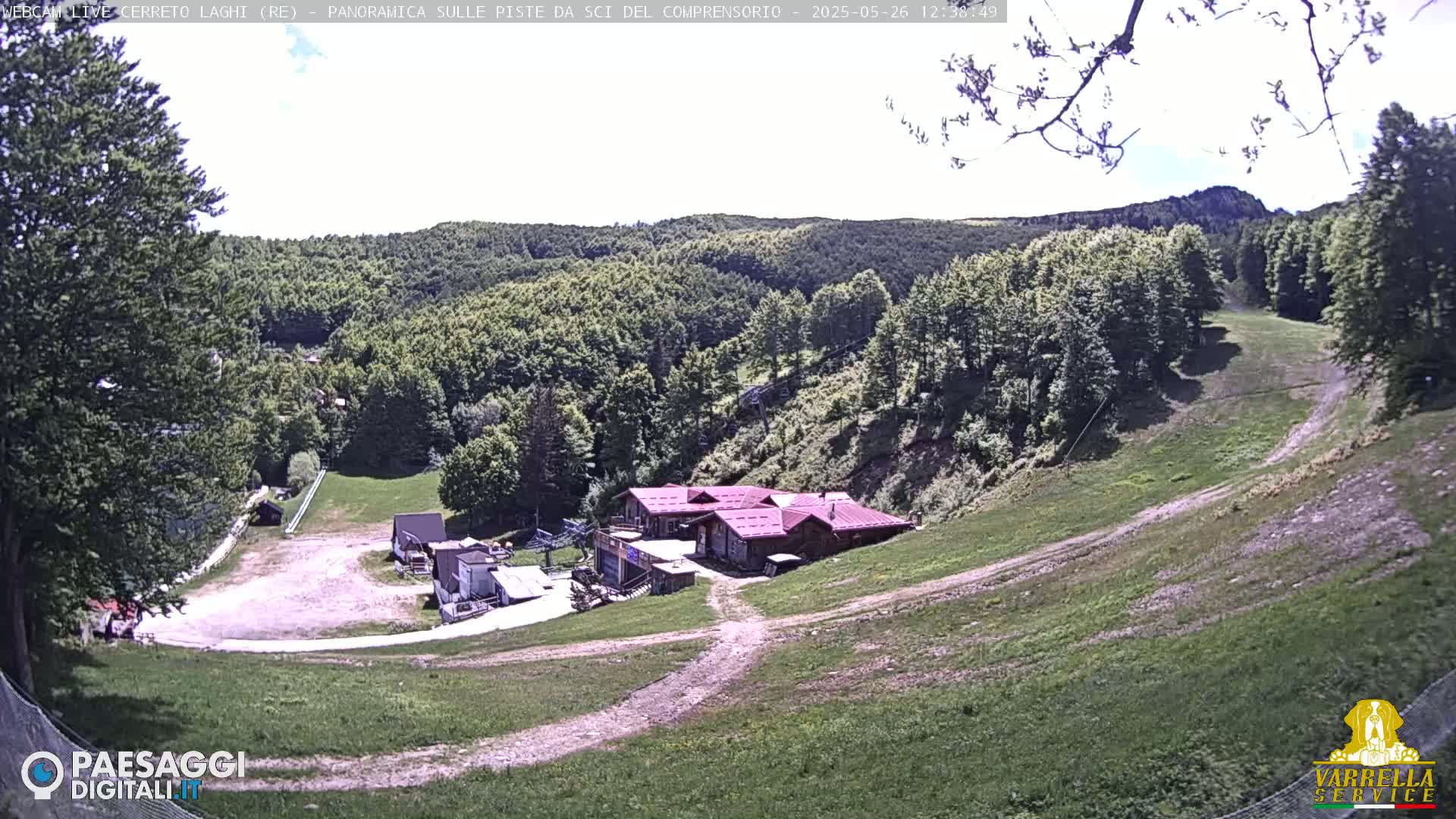 A mostly sunny view of a ski slope in a forested area, showing a building at the bottom of the hill and a mostly clear sky.