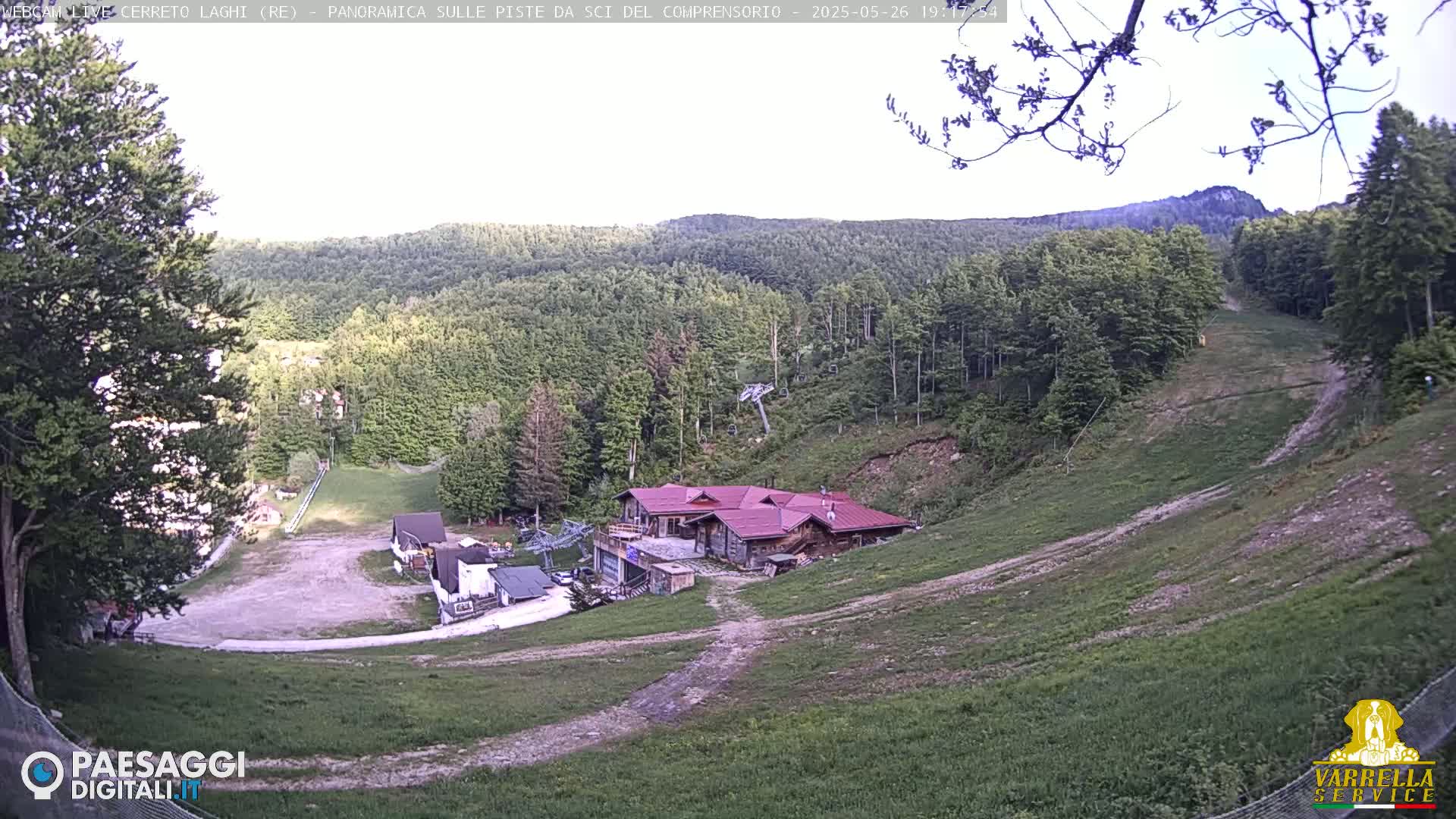 A mountain ski slope with a lodge and chairlift, surrounded by a lush green forest under a partly cloudy sky.
