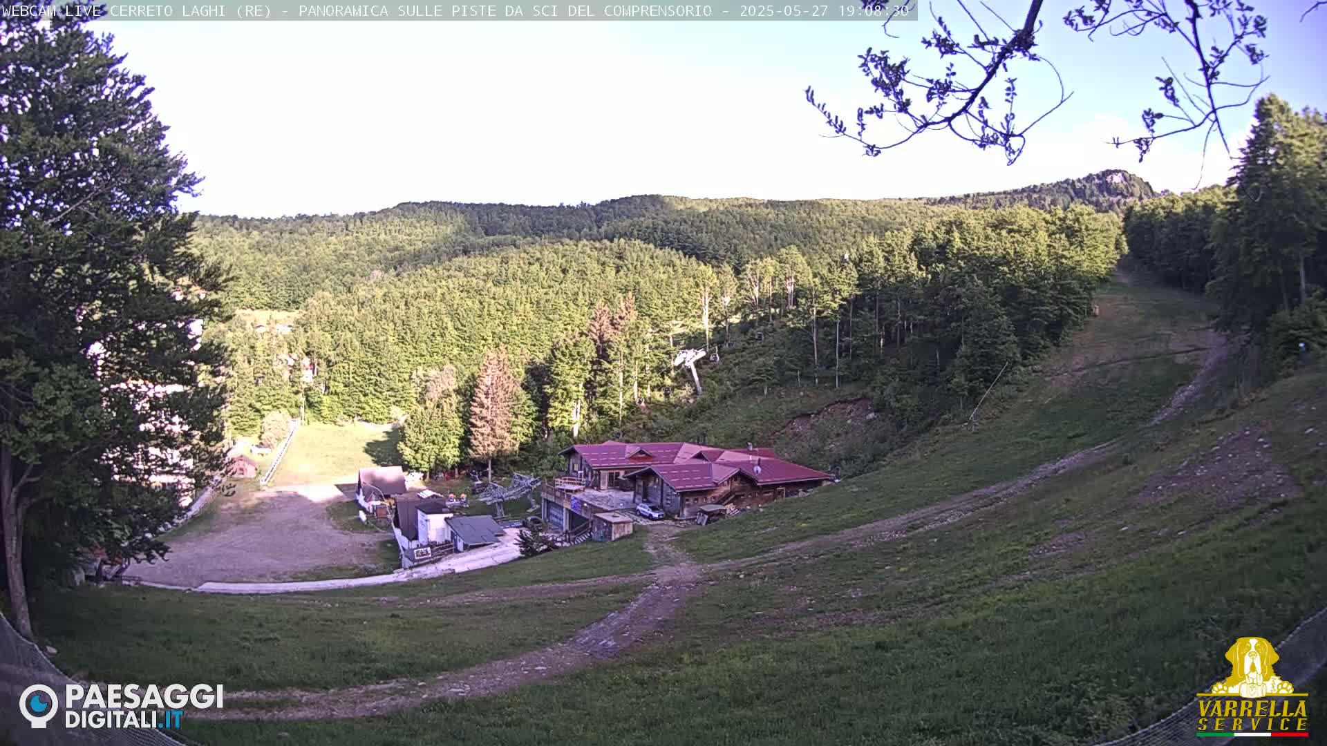 A mostly clear day reveals a mountainside ski slope with sparse vegetation, a cluster of buildings near the bottom, and a forest covering the upper slopes.