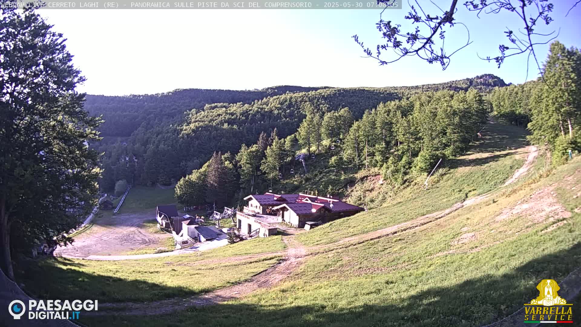 A mountainside ski resort, mostly green with some dirt paths, features a cluster of buildings near the bottom, under a mostly clear, sunny sky.