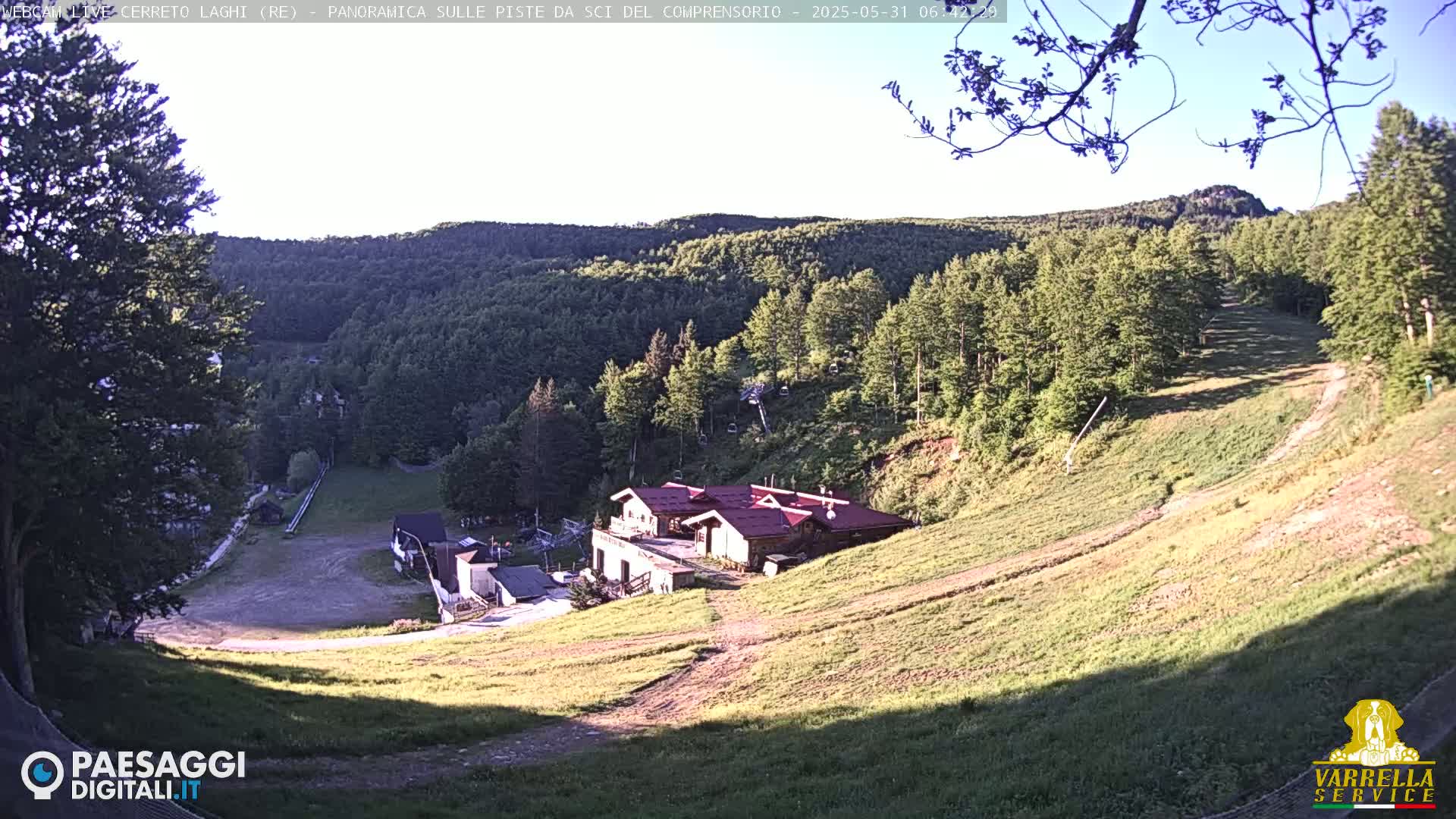 A grassy hillside slopes down towards a cluster of buildings nestled in a forest under a clear, sunny sky.