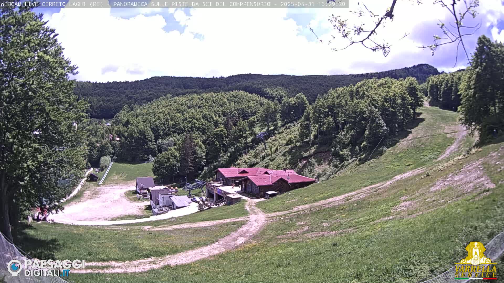 A partly cloudy day reveals a ski resort nestled in a green, hilly landscape, with several buildings and trails visible.
