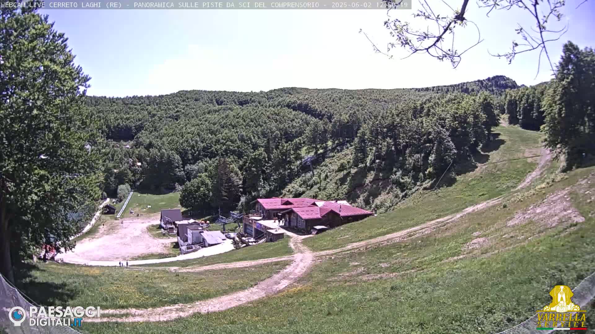 A mostly sunny view of a ski resort in the summer, showing grassy slopes, a small building complex, and a forested hillside in the background.