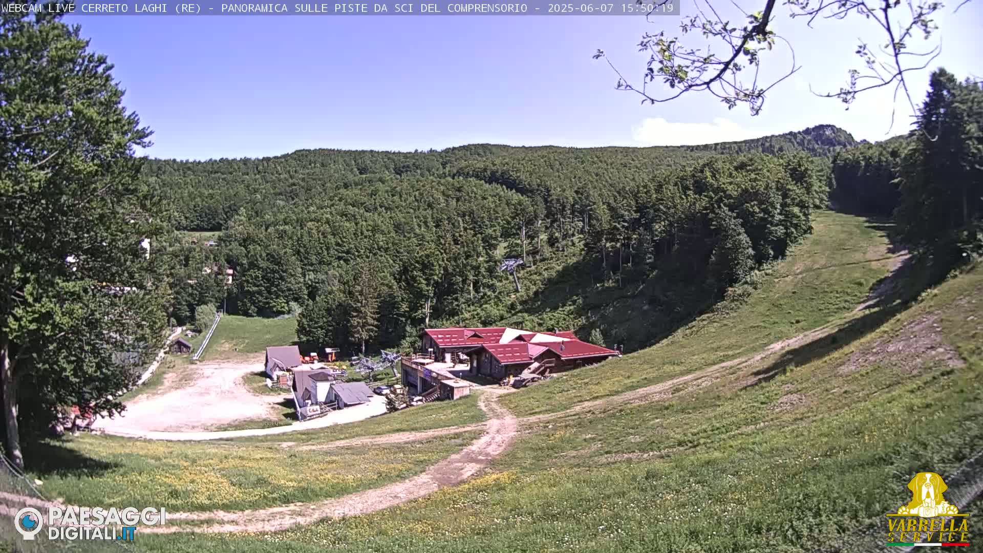 A mostly sunny view of a ski resort in the summer, showing a grassy hillside with a small building complex and a partially visible ski lift.