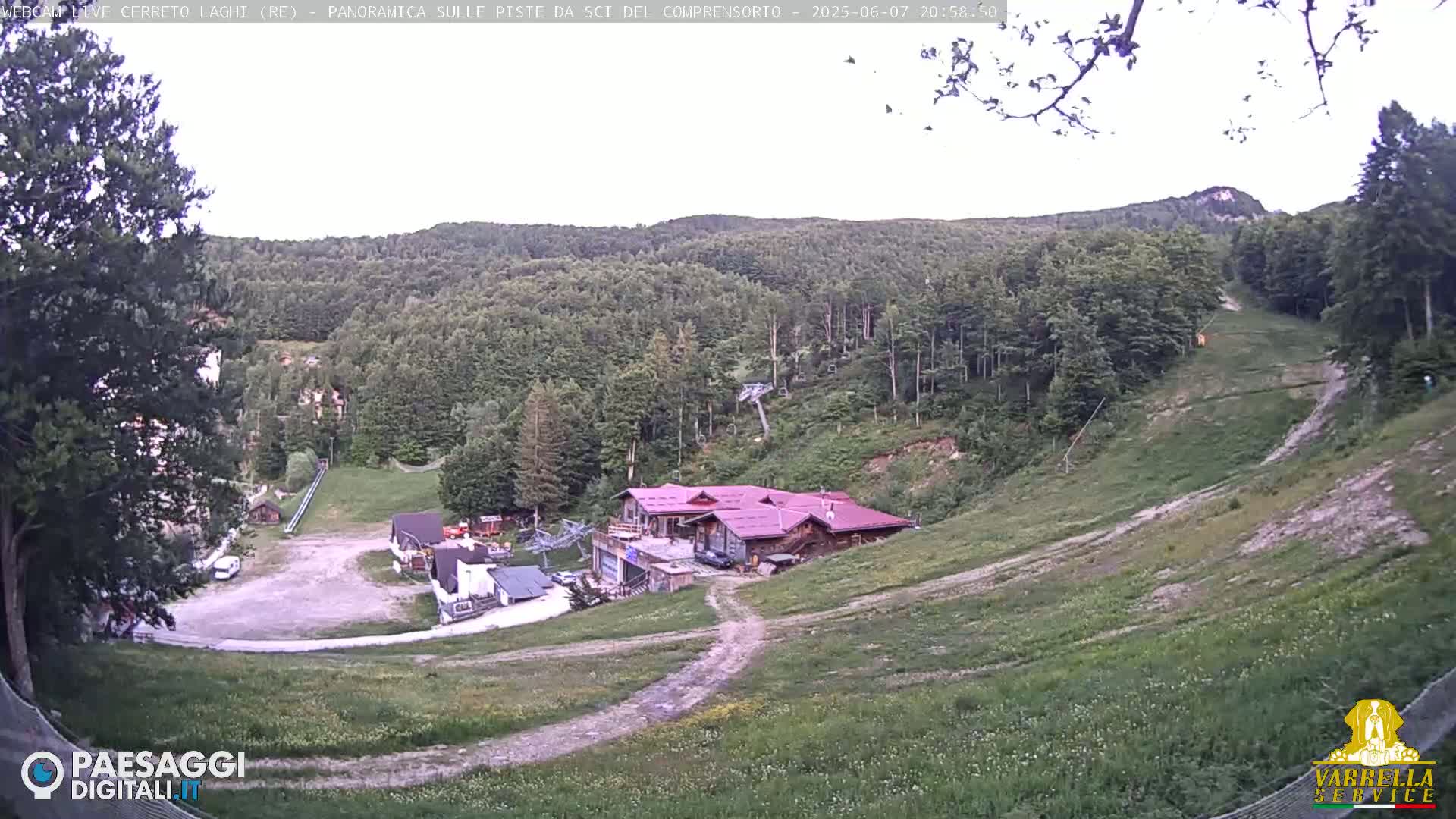 A mountainside ski resort, mostly green and grassy in the summer, with a few buildings and a chairlift visible under a mostly clear sky.